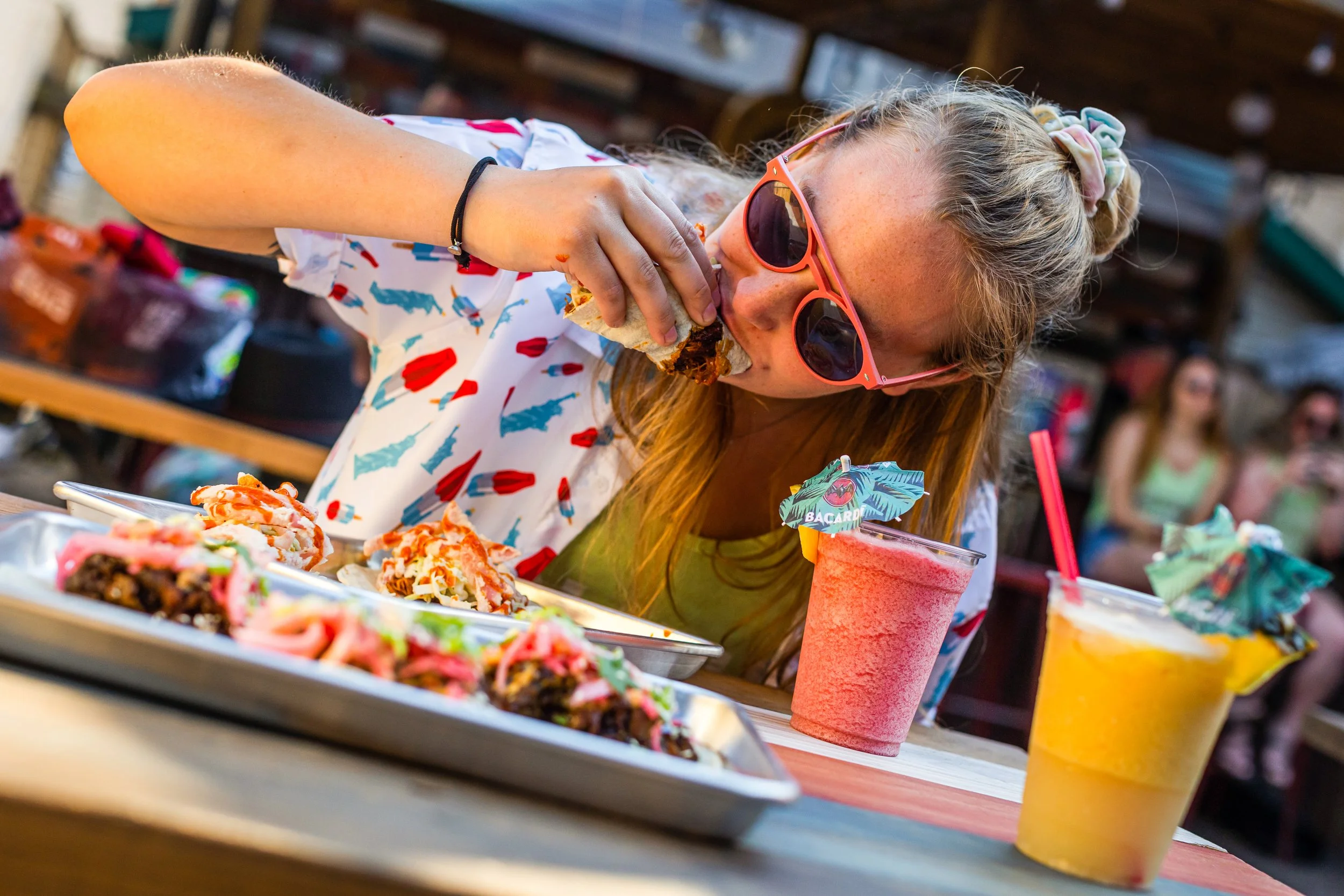 A woman in sunglasses and a patterned shirt eats tacos and drinks tropical smoothies at an outdoor table during daytime.