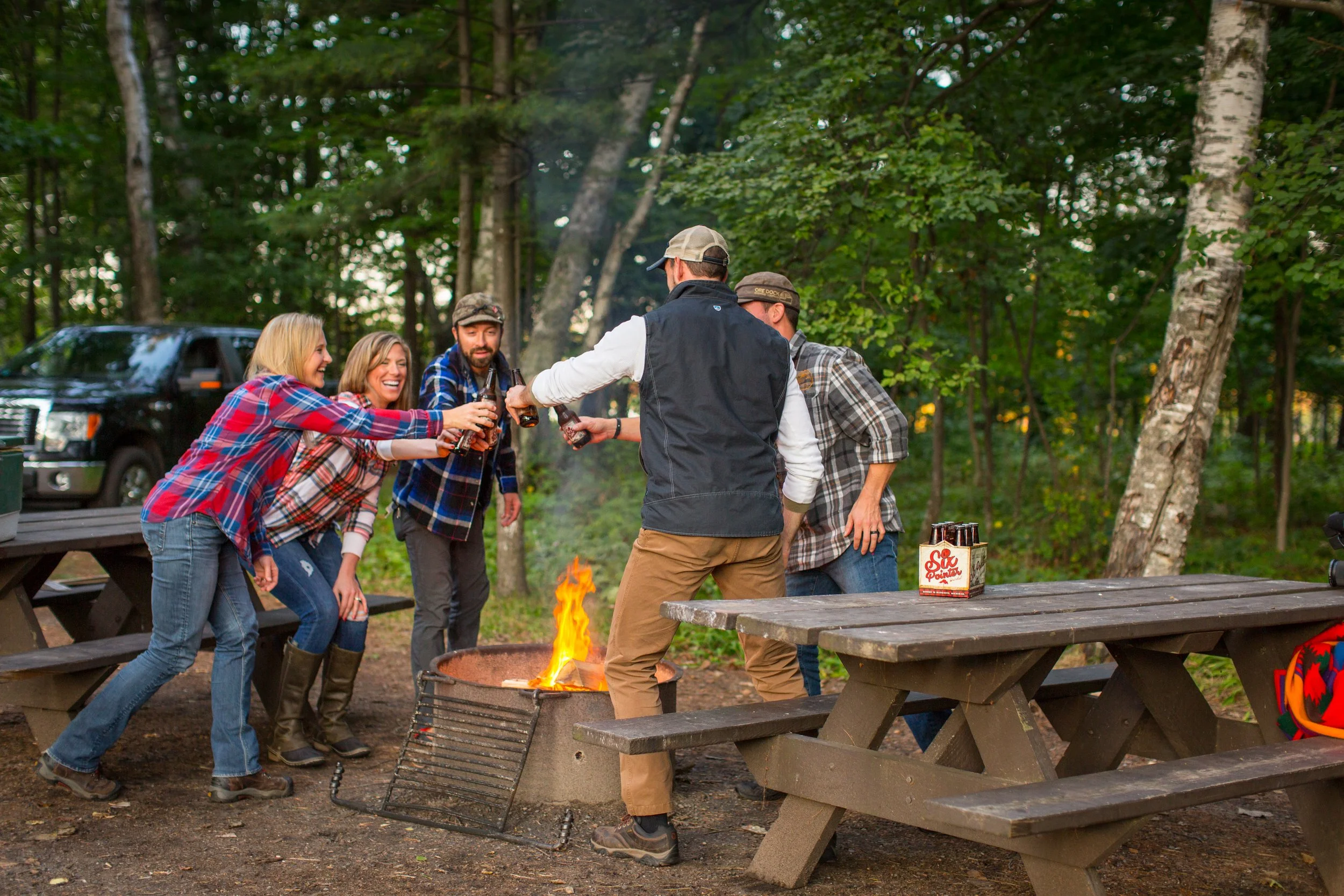 Group of people enjoying a campfire in a forested area, raising beer bottles in a toast at outdoor picnic tables.