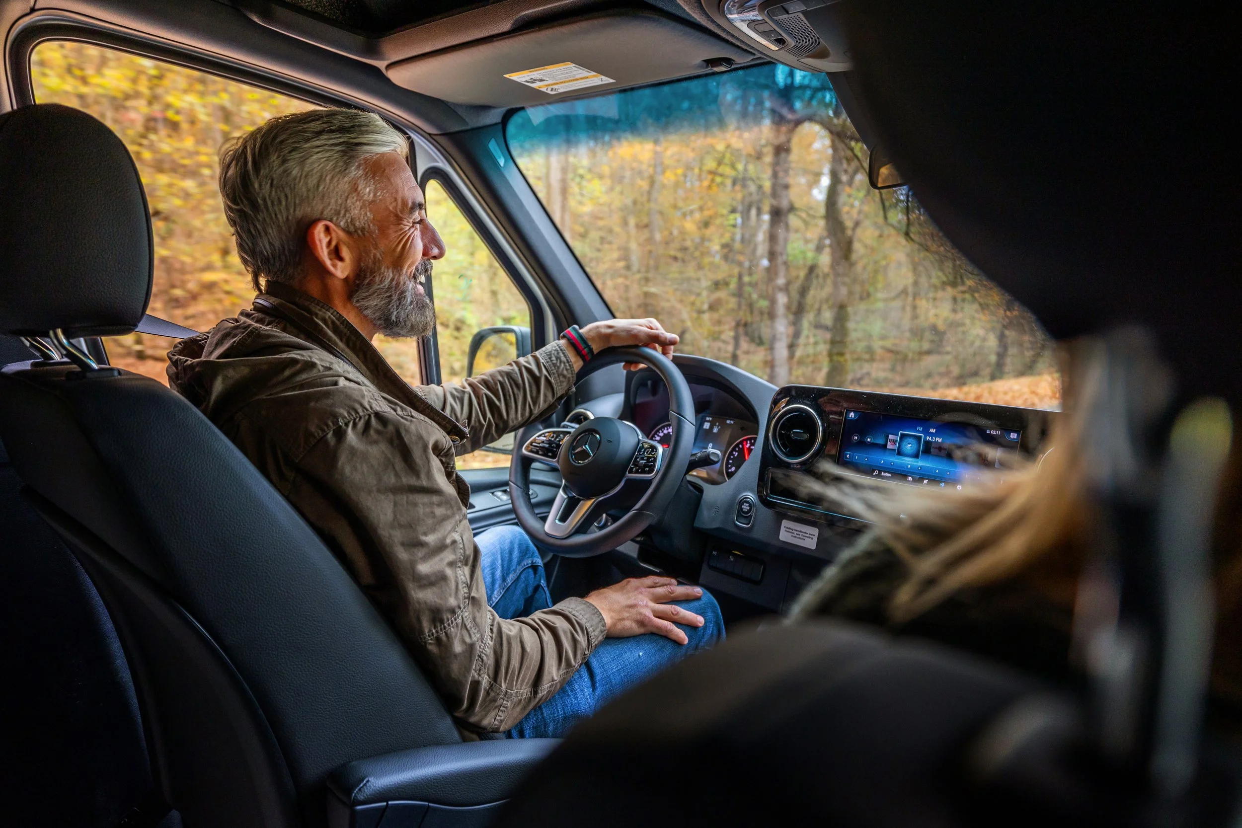 A man with gray hair and beard smiling and driving a Mercedes-Benz van through a wooded area with autumn foliage.