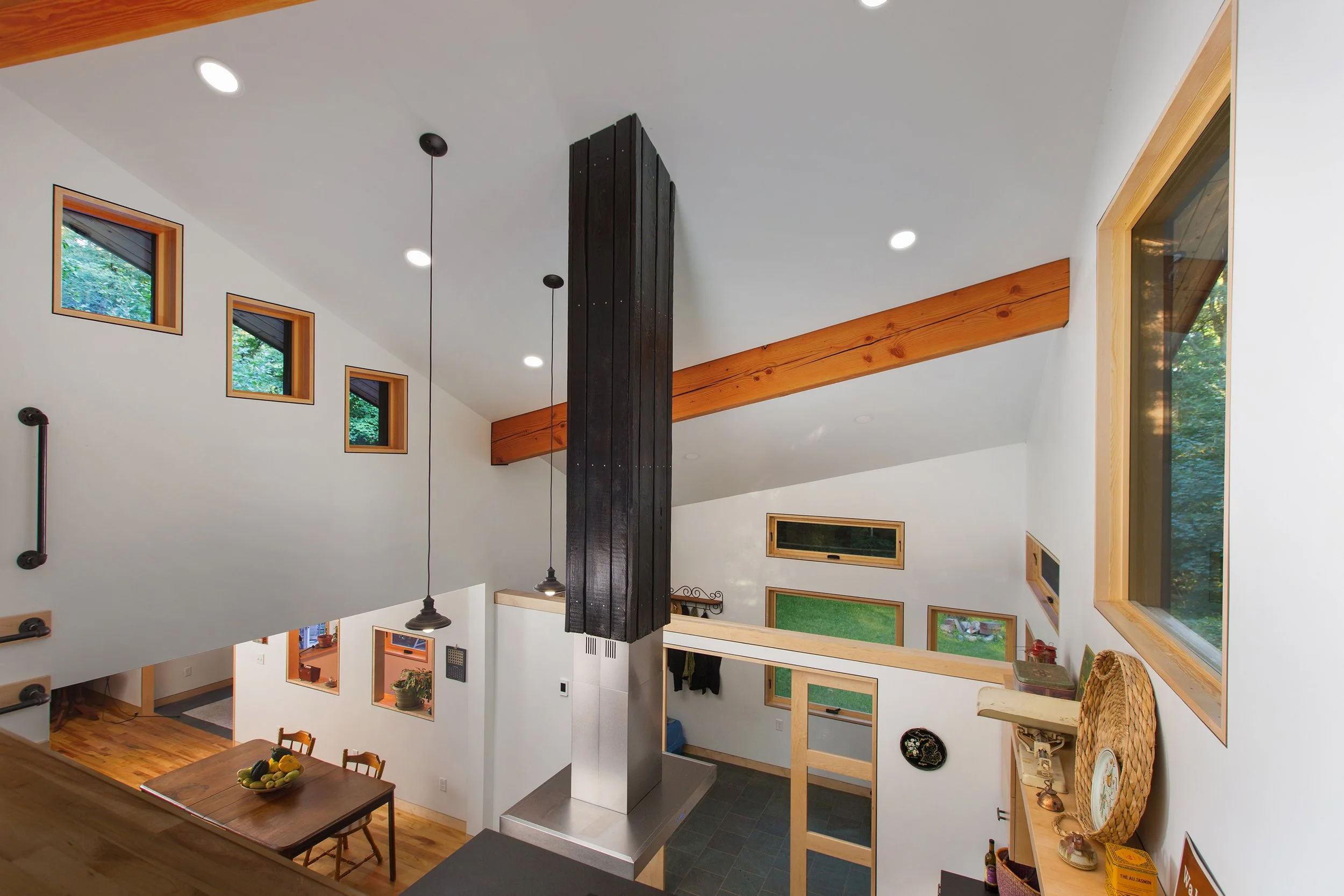 Interior view of a modern house with white walls, multiple small square and rectangular windows, wooden beams, and hanging pendant lights. A wooden dining table with chairs is visible on the lower level.