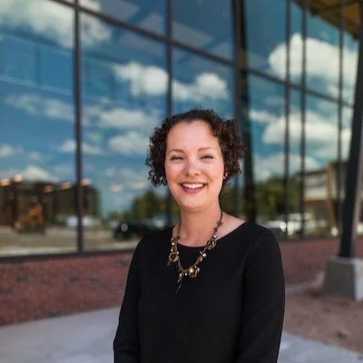 A smiling woman with curly hair wearing a black top and necklace stands outside a modern glass building with a blue sky and clouds reflected in the windows.