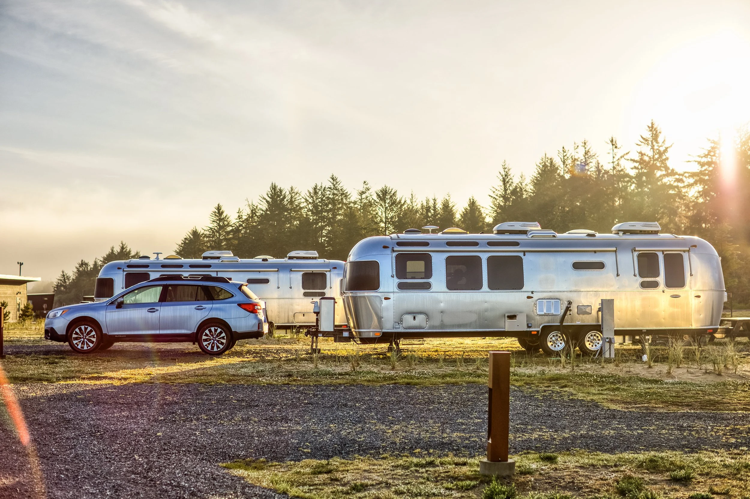 A silver SUV parked beside two shiny aluminum travel trailers in a grassy outdoor area with trees in the background and the sun shining brightly.