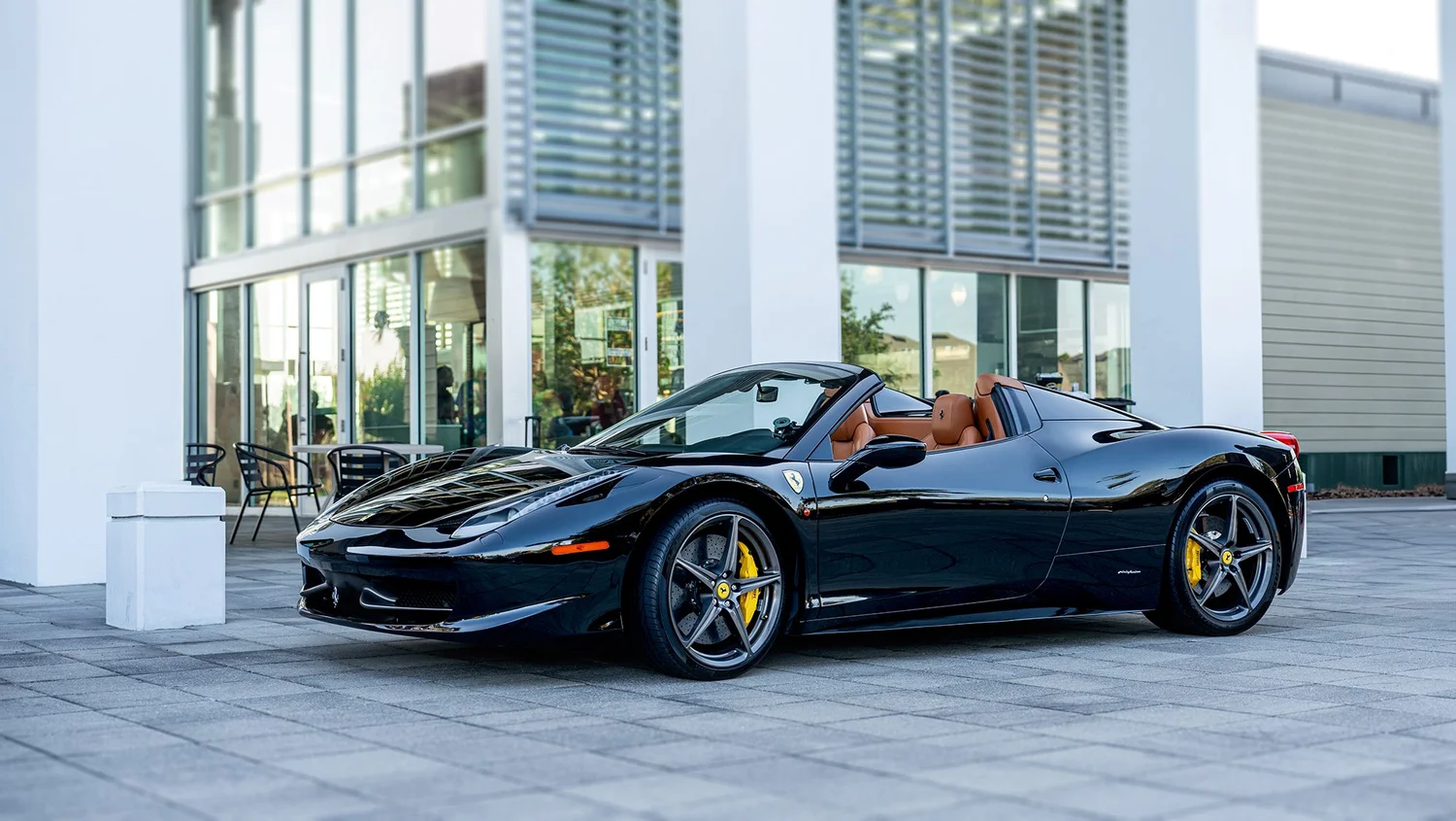 Black Ferrari convertible with tan interior parked outside a modern building with large glass windows and a patio area with chairs.