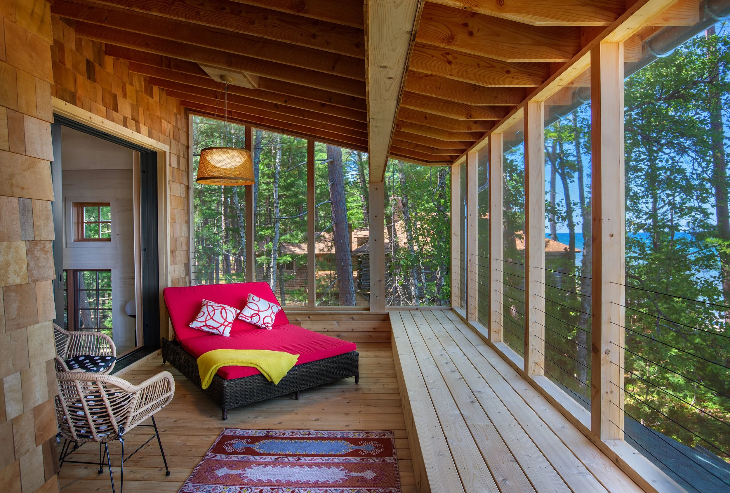 Cozy screened-in porch with wooden flooring, a black wicker sofa with pink cushions and pillows, wicker chairs, a patterned rug, surrounded by tall trees and a view of a lake.