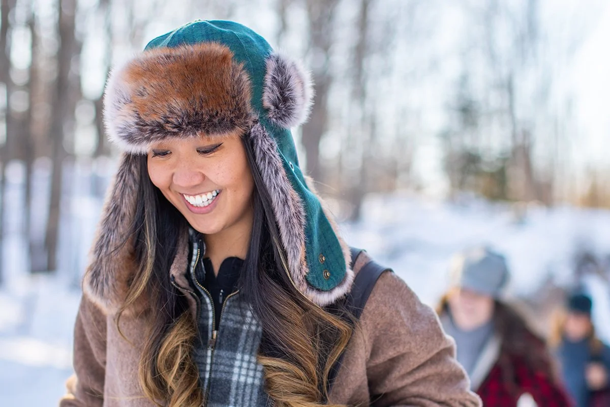 A woman wearing a fur-lined winter hat and a brown coat, smiling outdoors in a snowy forest with children in the background.