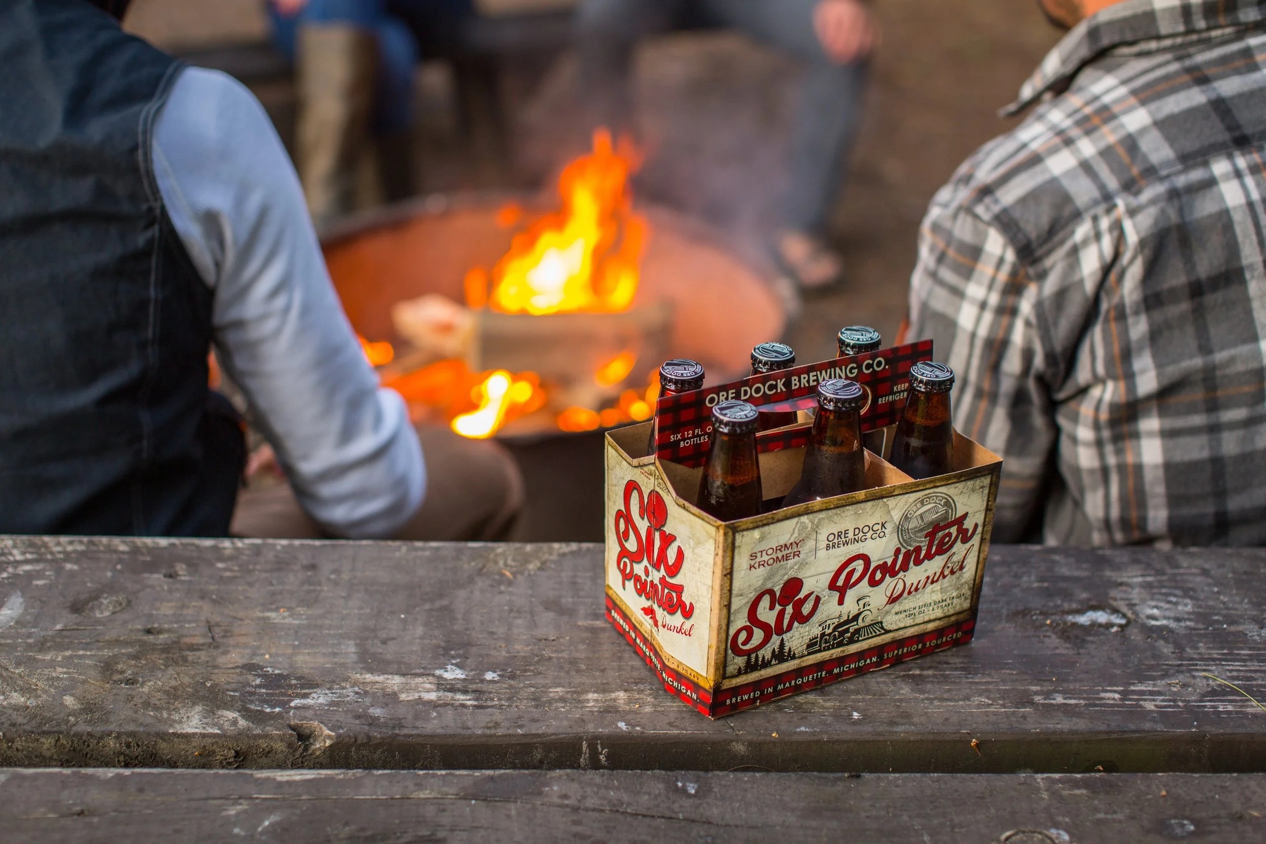 A six-pack of Six Pointer Dunkel beer bottles on a weathered wooden picnic table, with a bonfire in the background and two people sitting nearby.