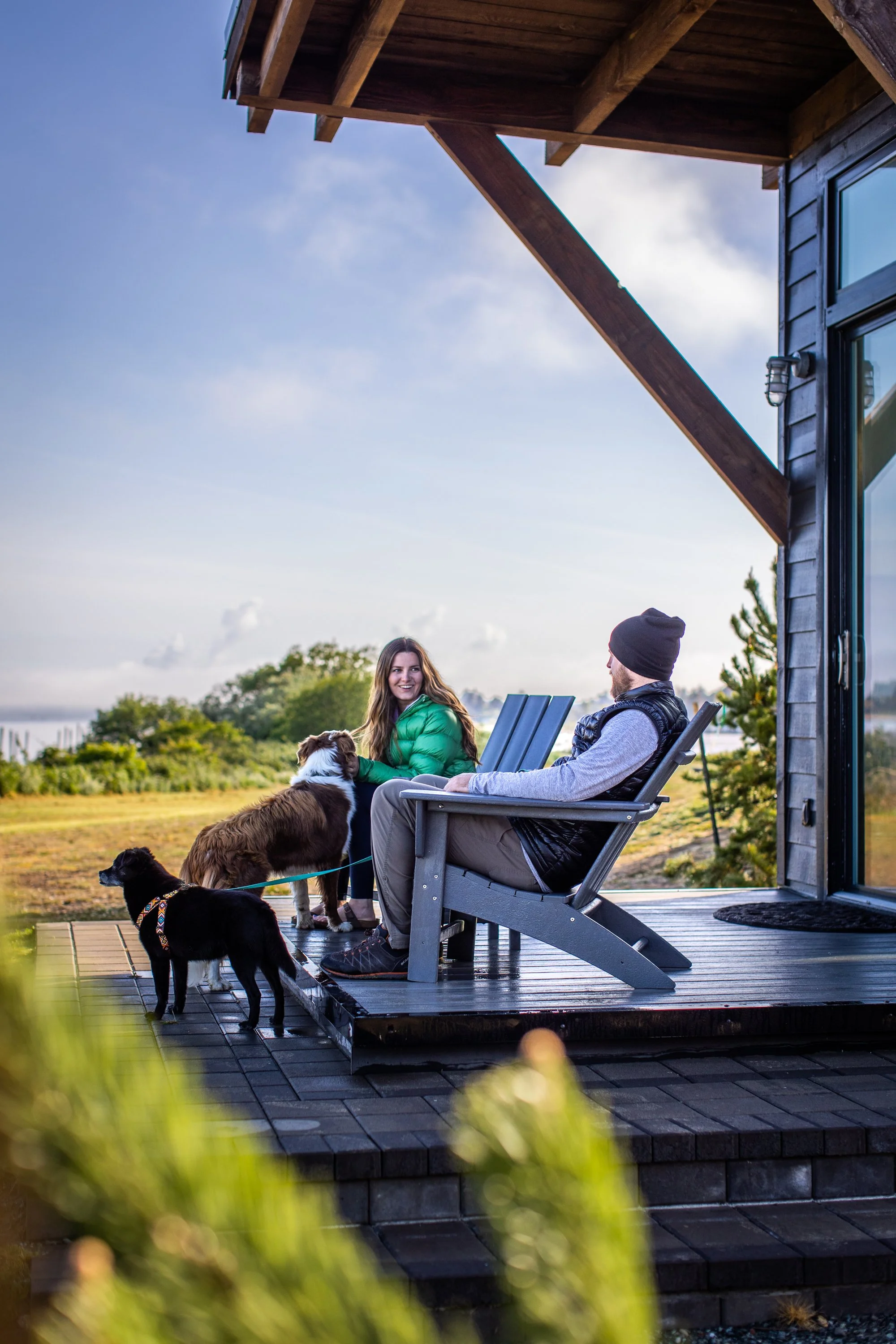 A man and woman sitting on a wooden front porch with three dogs, enjoying a conversation outdoors during daytime.
