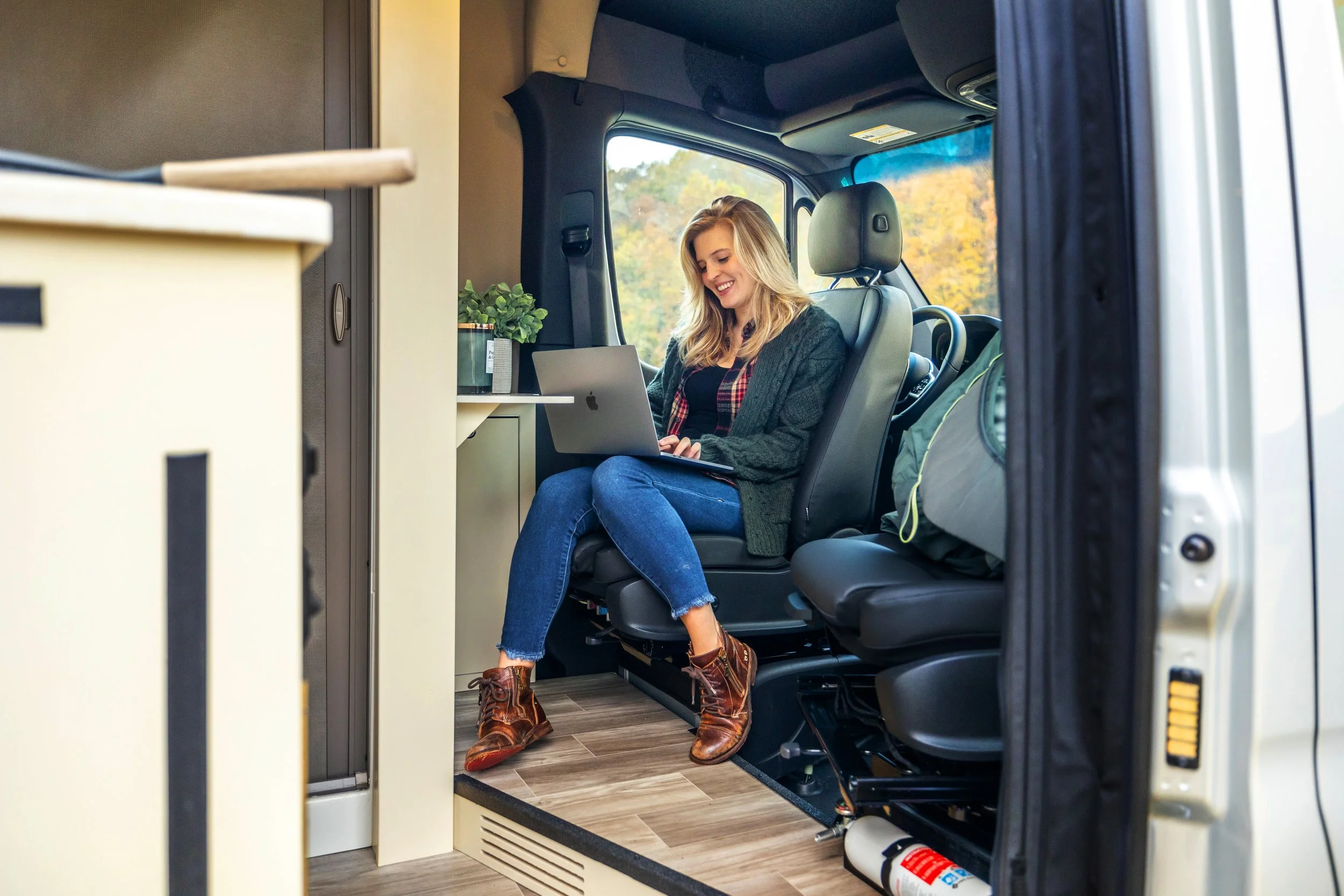 A young woman sitting inside a camper van, working on a laptop and smiling, with travel gear visible behind her.