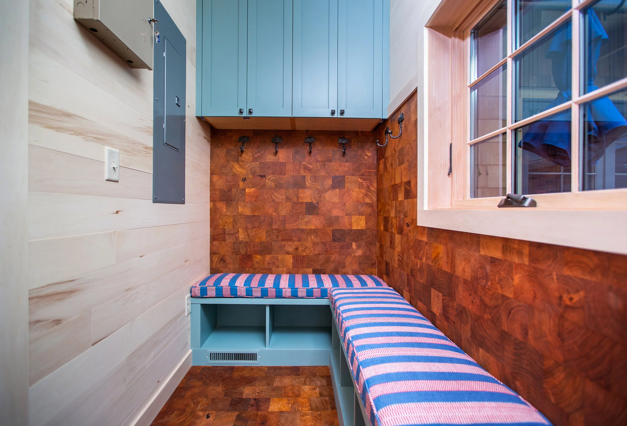 Small room with a built-in bench with a striped cushion, a window, a blue cabinet, and hooks on a wood-paneled wall.