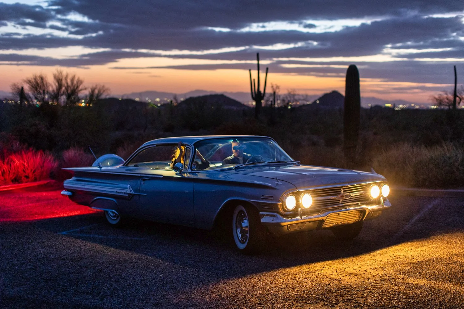 A vintage blue car with headlights on parked on a desert road at sunset, with cacti and desert shrubs in the background.