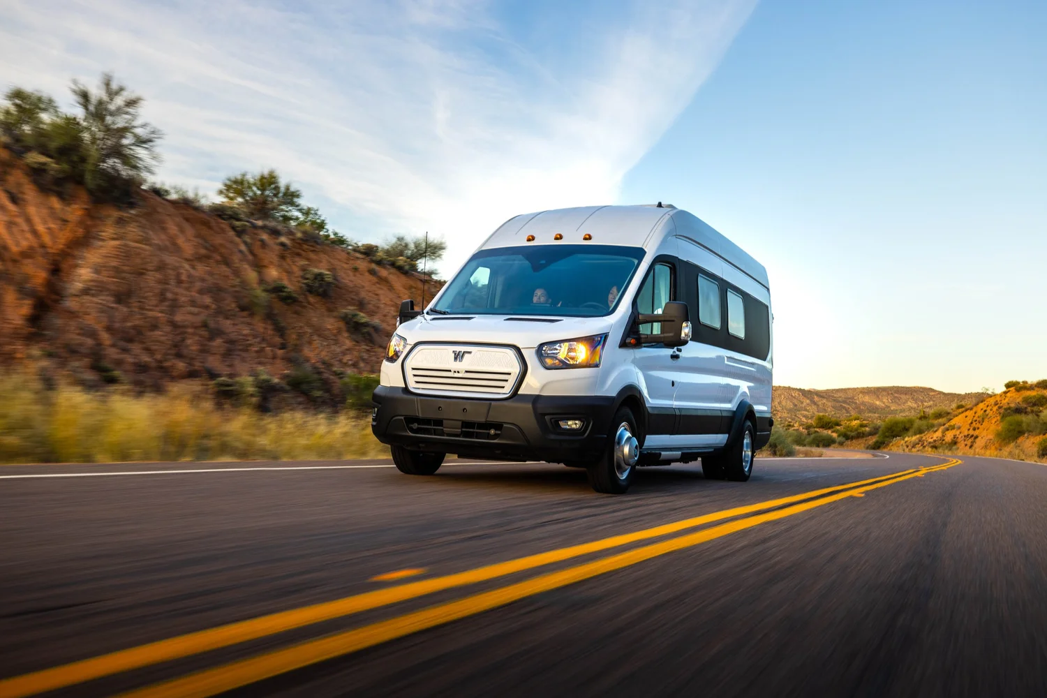 A white camper van driving on a winding road through a desert landscape with red rock formations and sparse vegetation, under a partly cloudy sky.