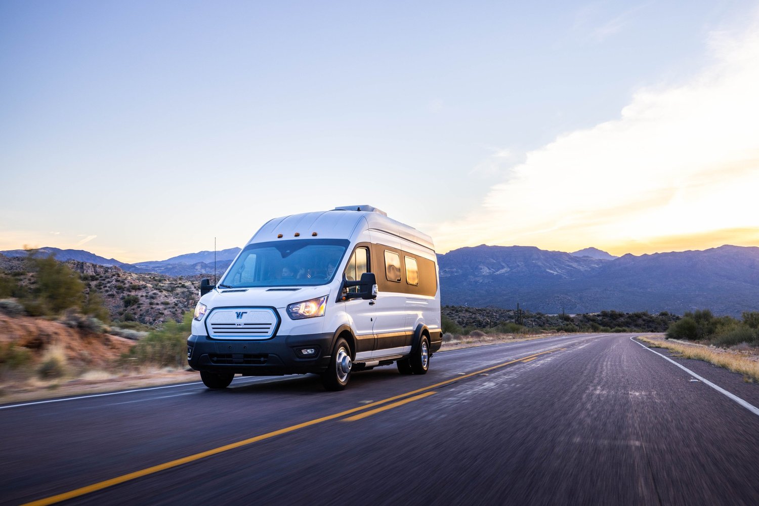 A white camper van driving on a two-lane road through a desert landscape with mountains in the background at sunset.