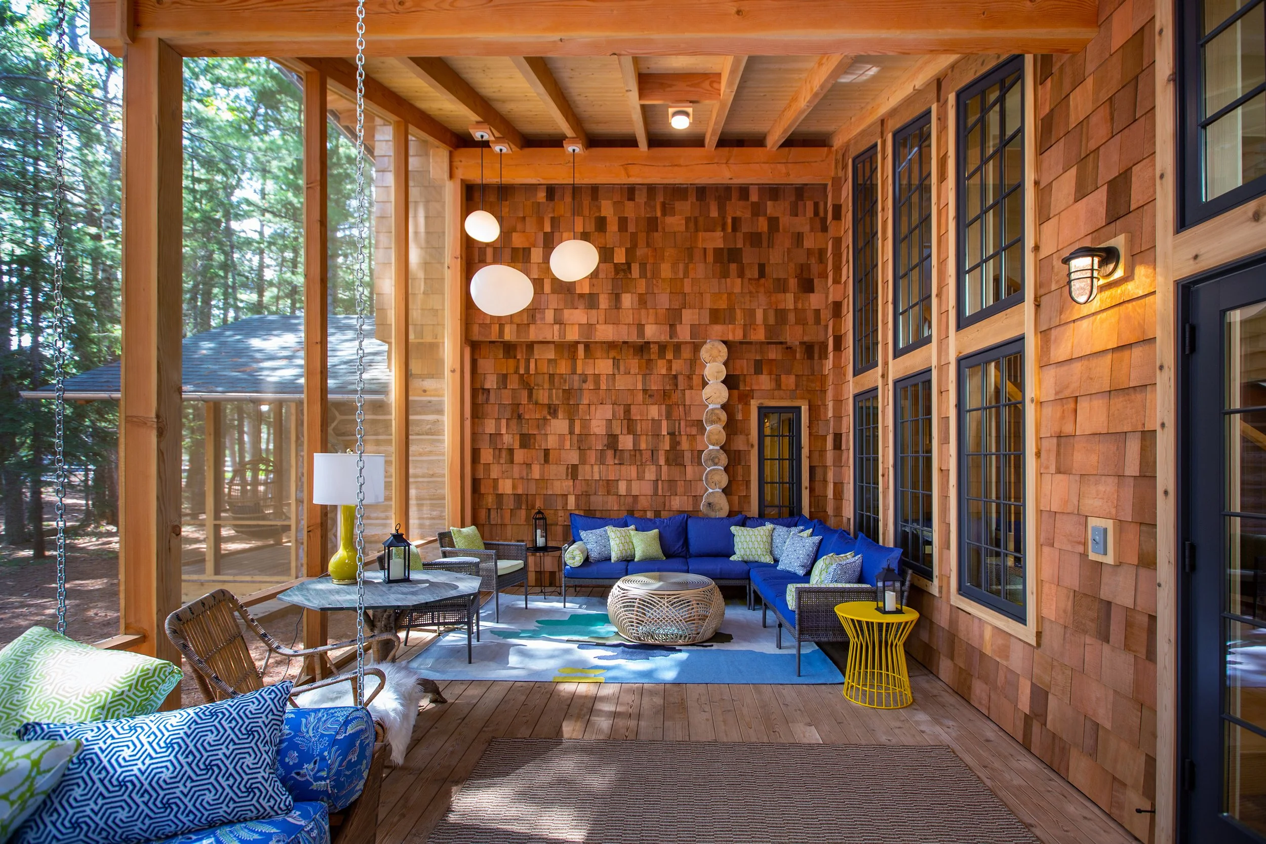 A screened porch with wooden walls and ceiling, outdoor furniture including a sectional sofa with blue cushions, a wicker chair with patterned pillows, a small yellow side table, and a round woven coffee table. The porch has large windows, hanging pe