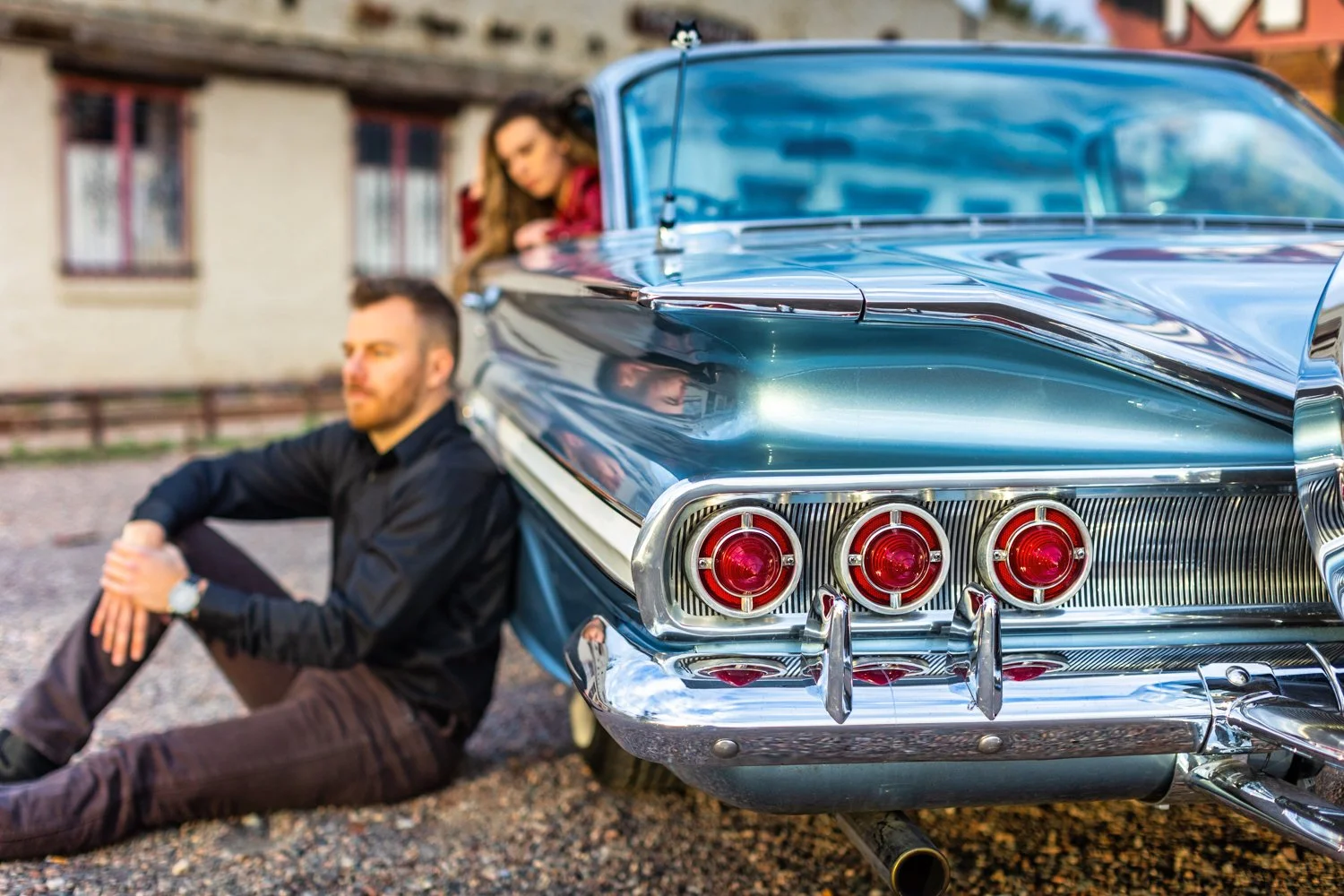 A young man with a beard and dark shirt sitting on the ground beside a classic 1960s blue car, with a woman inside the car leaning out to look at him. The car has chrome details and round taillights, parked in front of a rustic building.