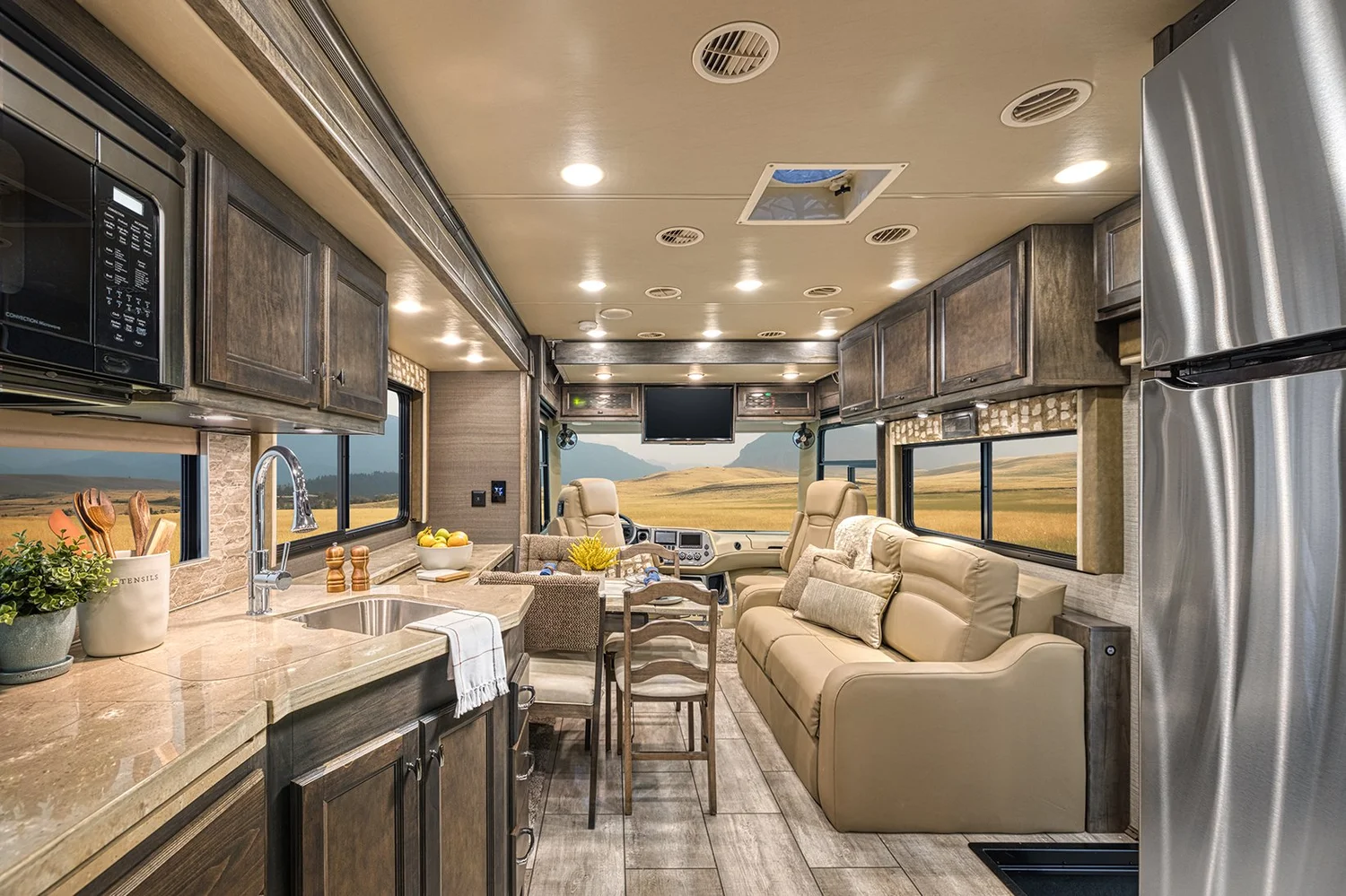 Interior of a modern RV with a kitchen area, dining table, and beige seating, large windows showing open landscape, and an overhead TV.