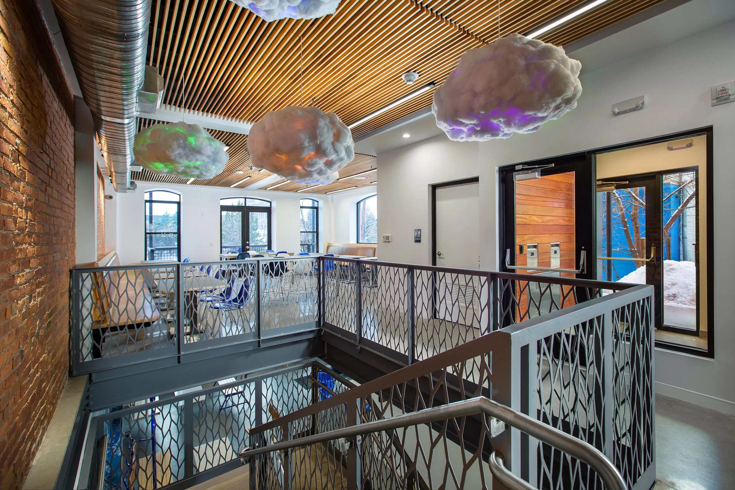 Interior view of a modern building with decorative cloud-shaped light fixtures hanging from a wooden slat ceiling, large windows, a brick accent wall, and a staircase with geometric-patterned railing.