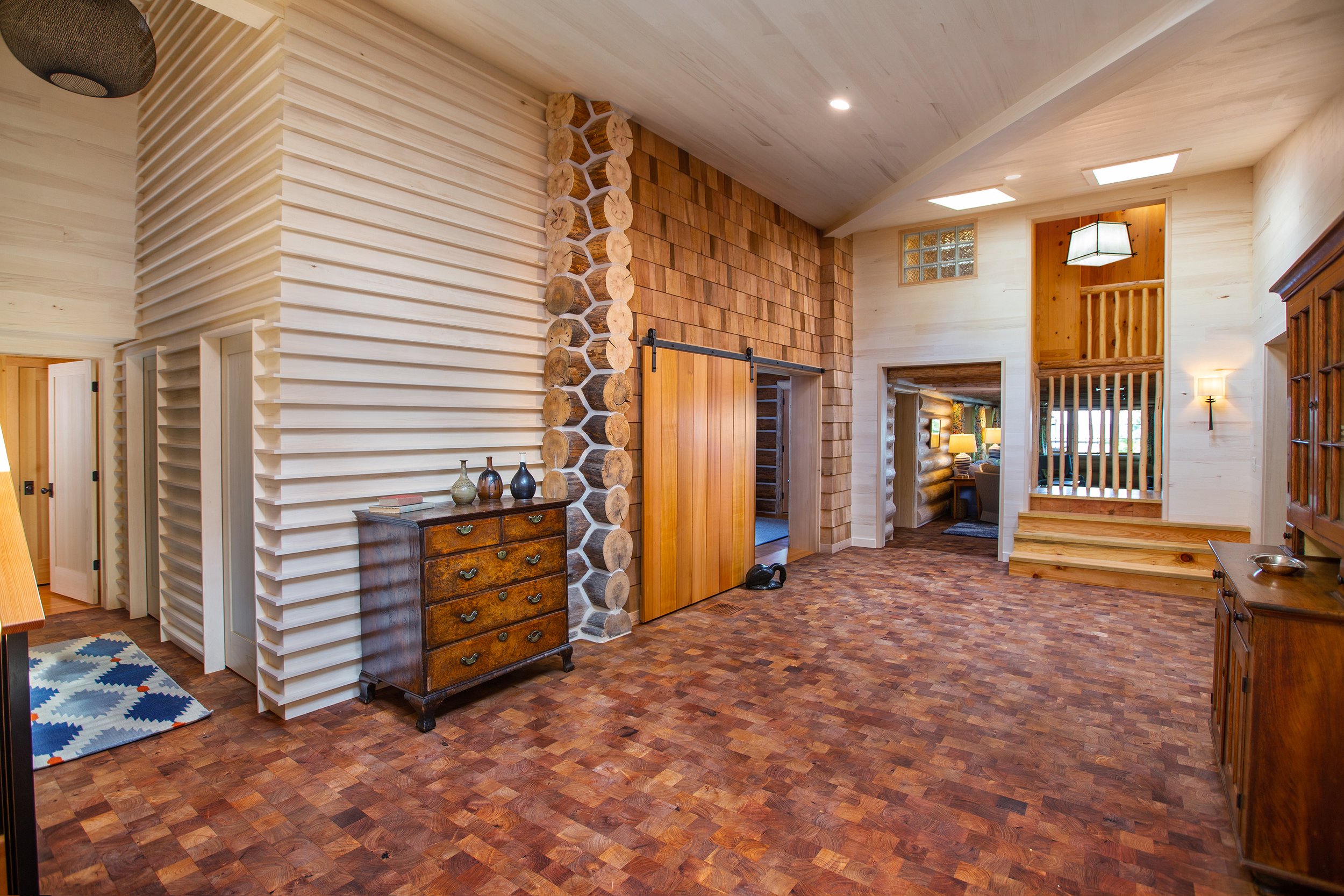 Interior of a rustic-style living room with wood paneling, log accents, hardwood flooring, and wooden furniture, including a small dresser with decorative bottles on top.
