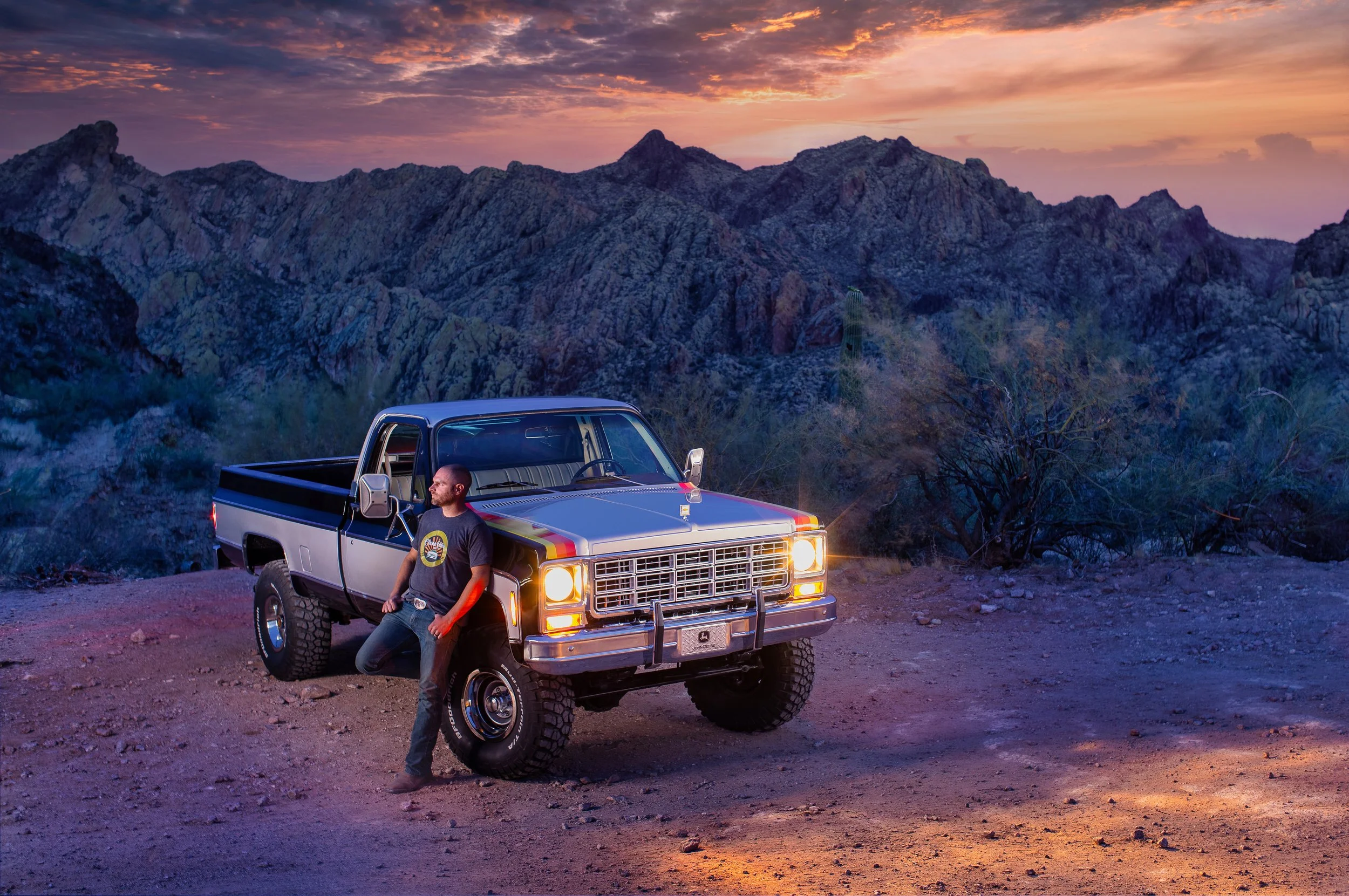 A man leaning against a pickup truck with mountains and a colorful sunset in the background.