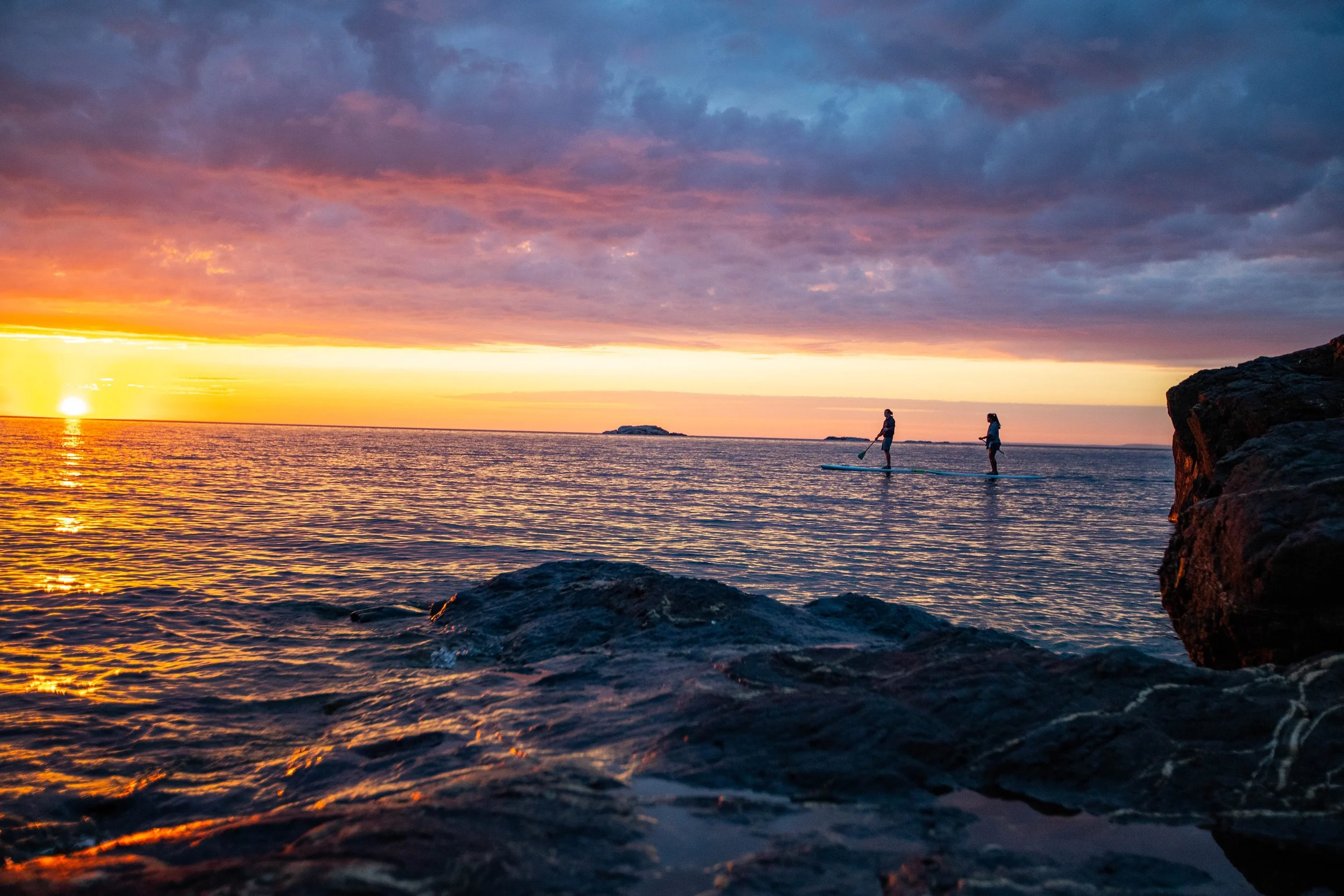 Two people stand on paddleboards on a calm ocean during sunset, with rocky shoreline in the foreground and an island in the distance.
