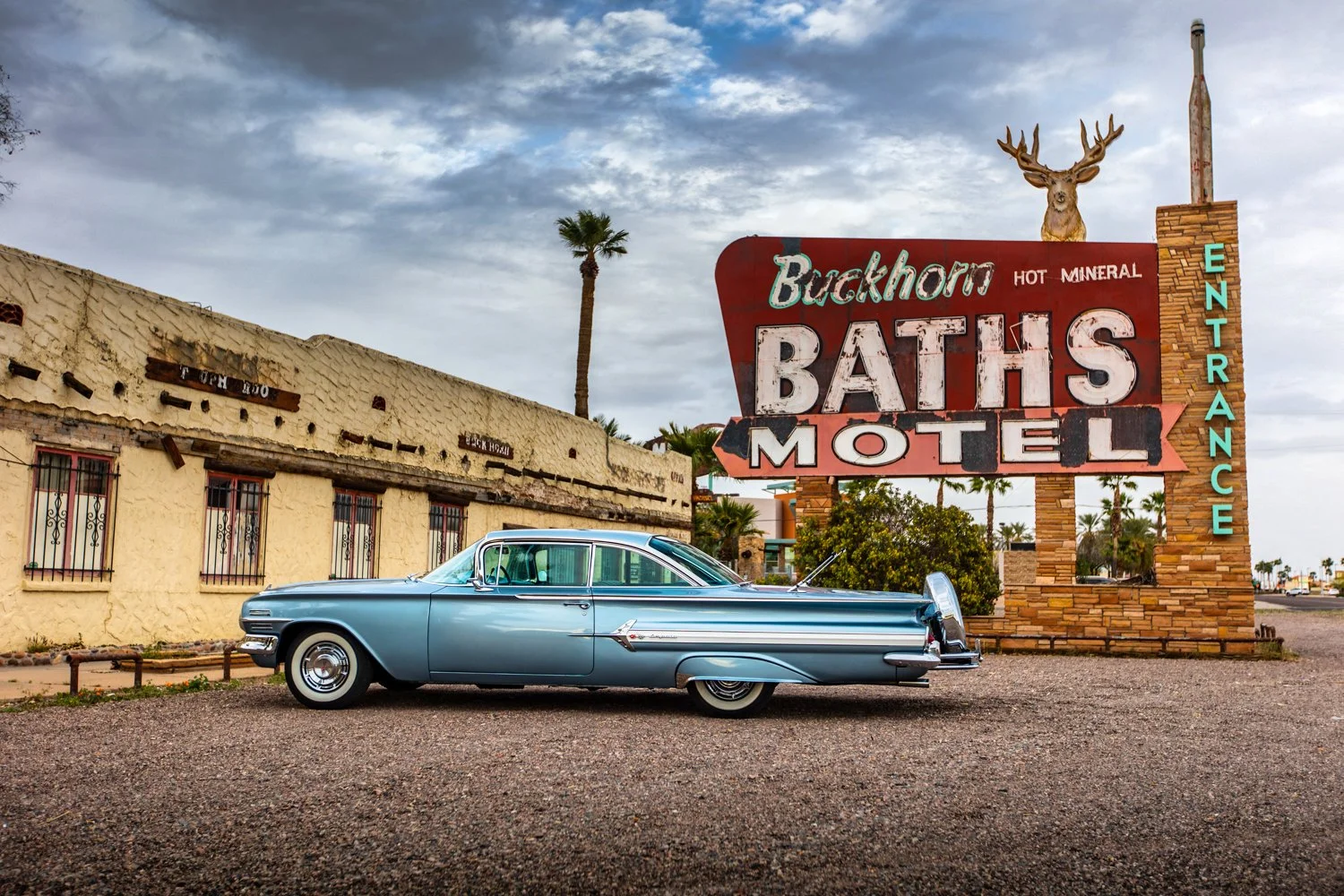 A vintage blue car parked in front of a retro sign for Buckhorn Baths Motel with a deer statue on top of the sign, palm trees, an old building, and a cloudy sky in the background.
