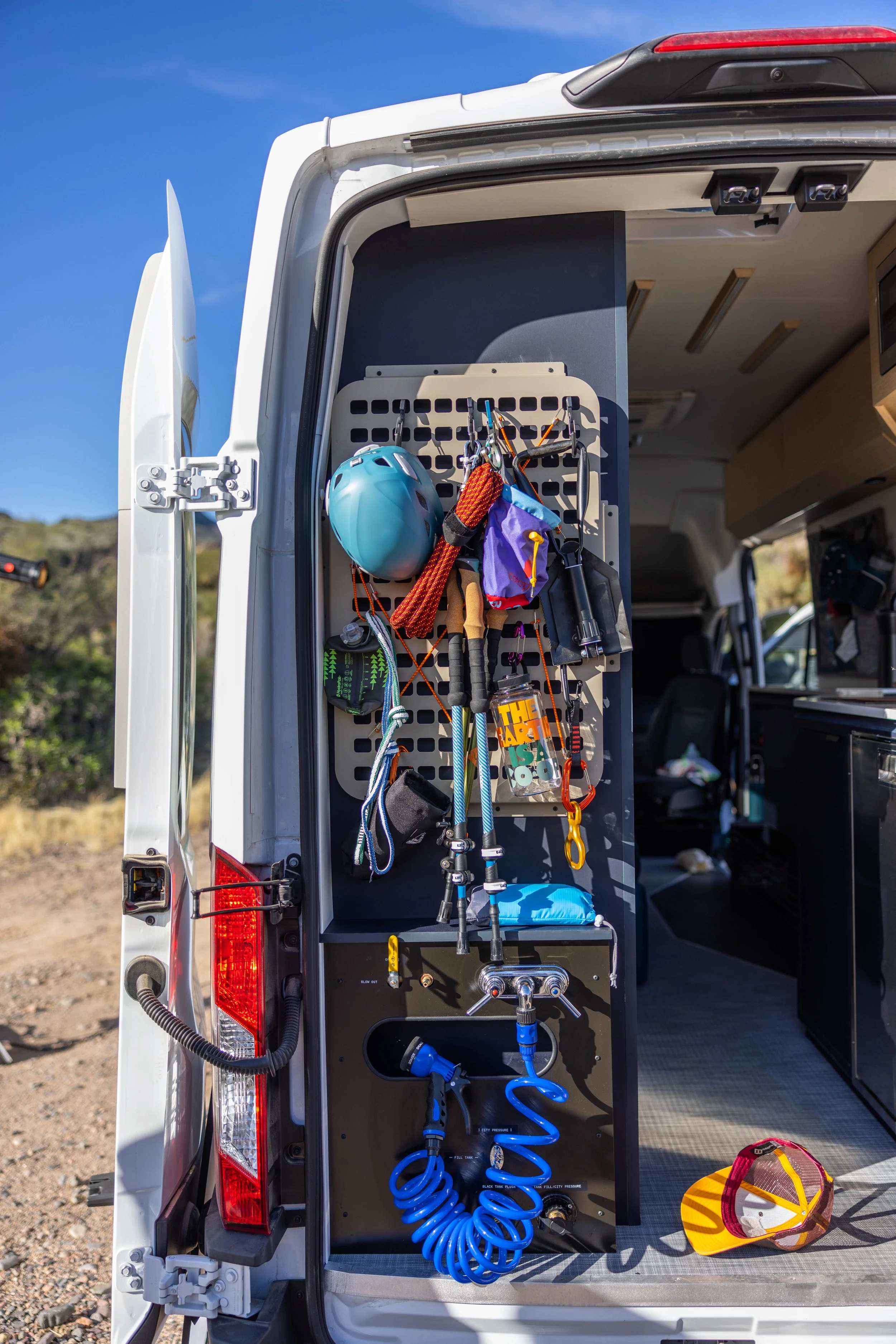 Open van showing climbing and outdoor gear, including a helmet, ropes, carabiners, a water hose, and other equipment hanging on hooks inside the vehicle, with a mountainous landscape in the background.