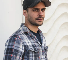 A young man with a beard and mustache wearing a gray cap and a plaid shirt, looking at the camera, standing against a white textured wall.