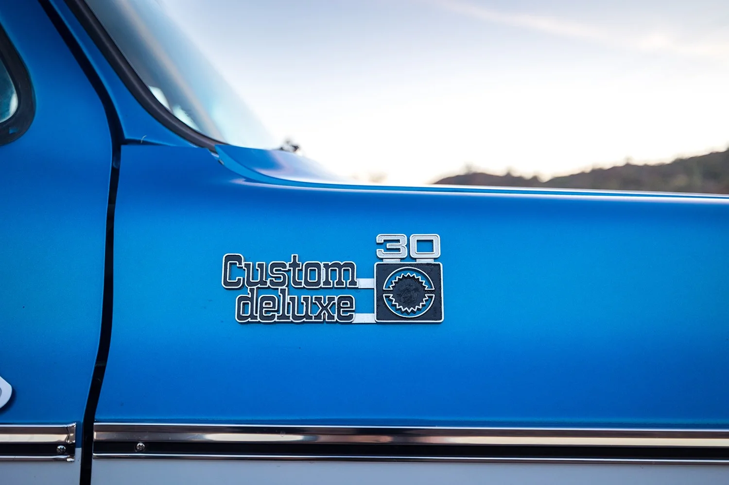 A close-up of the side of a vintage blue Chevrolet truck with a badge reading 'Custom Deluxe 30' and a gear shift knob graphic.