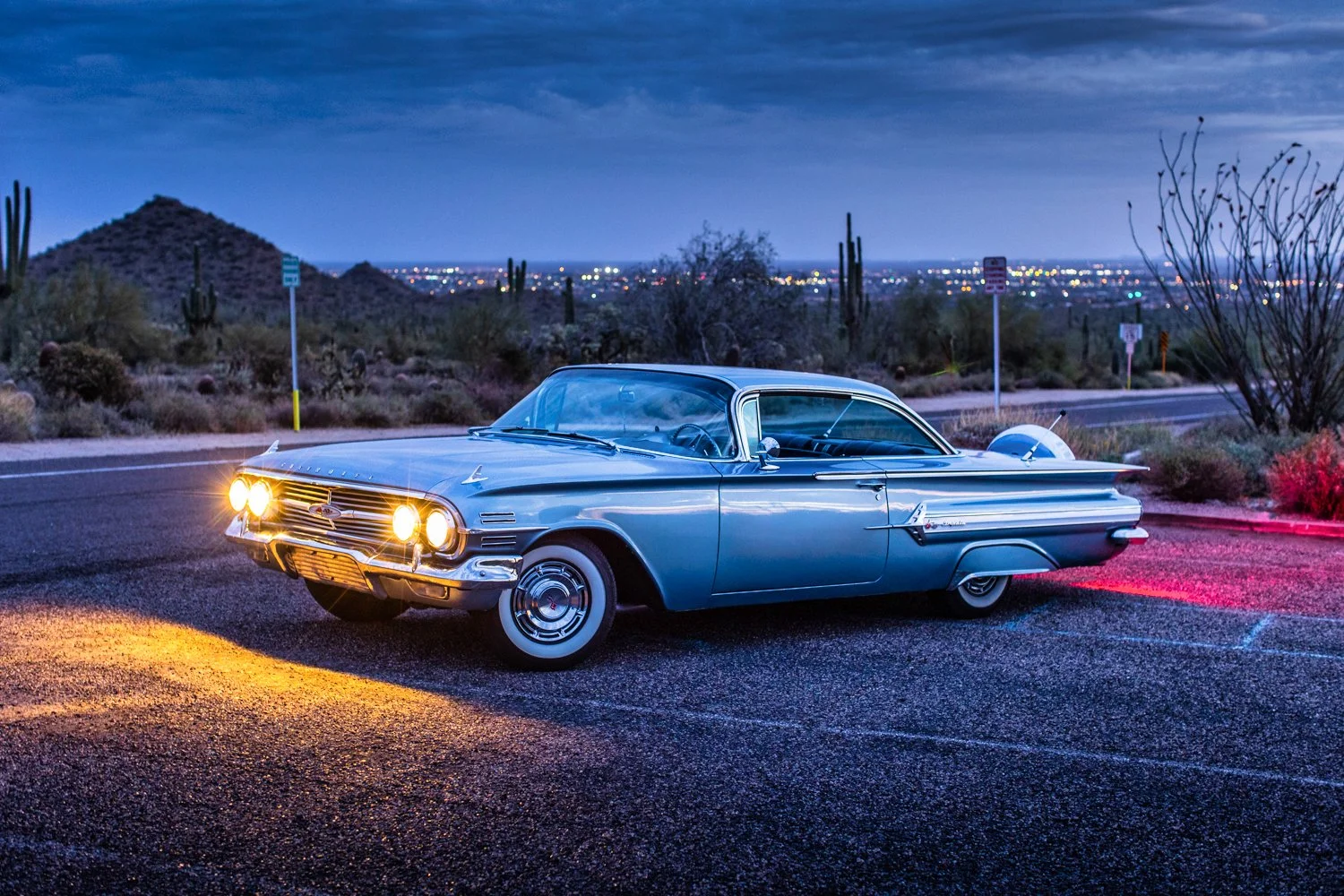 A vintage light blue classic car parked on a desert road at dusk with cacti and mountains in the background.