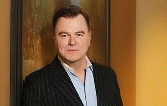 Portrait of a man in a black pinstripe suit and light blue shirt standing indoors with a warm-colored wall in the background.