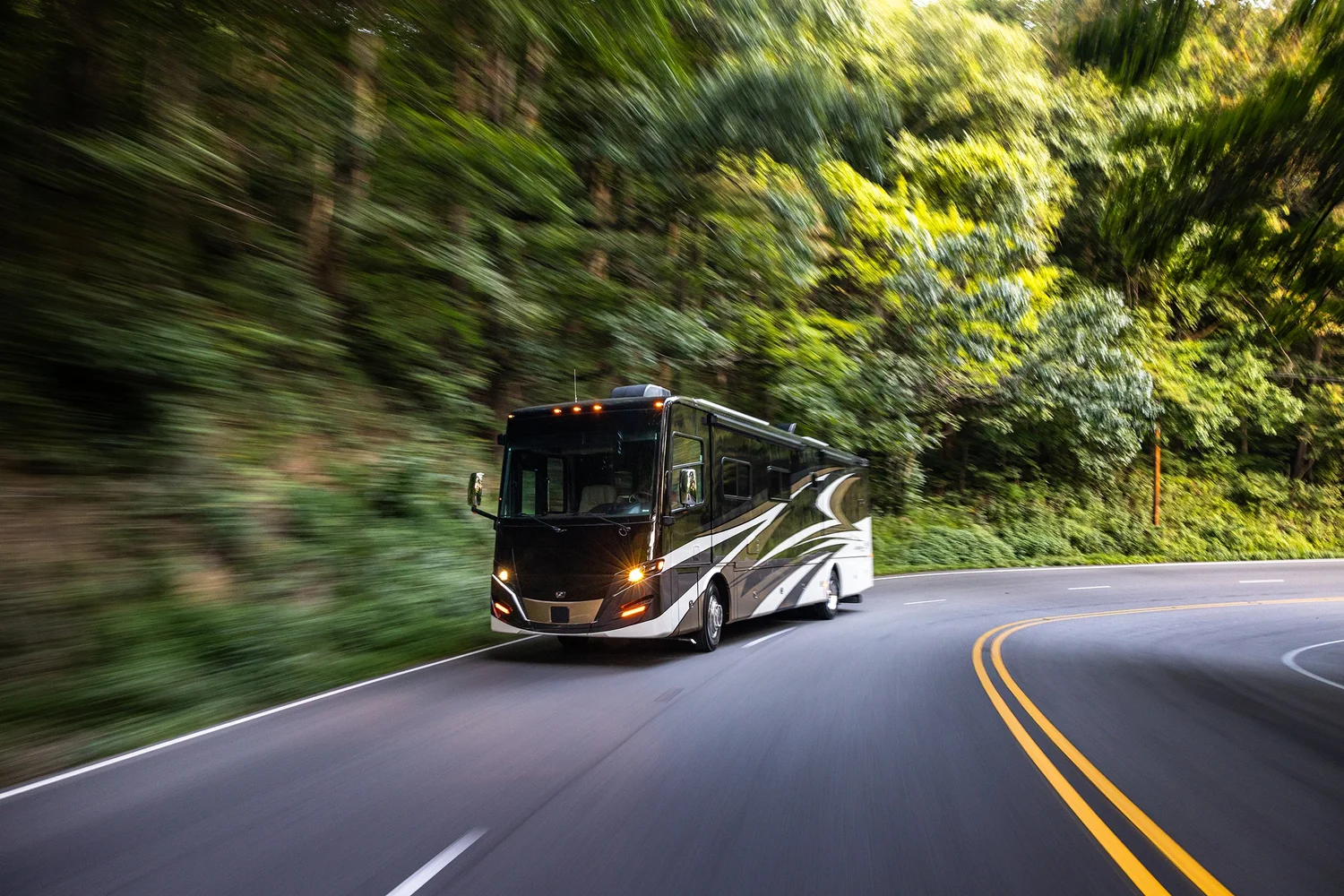 A black and white motorhome driving along a curving road surrounded by lush green trees.