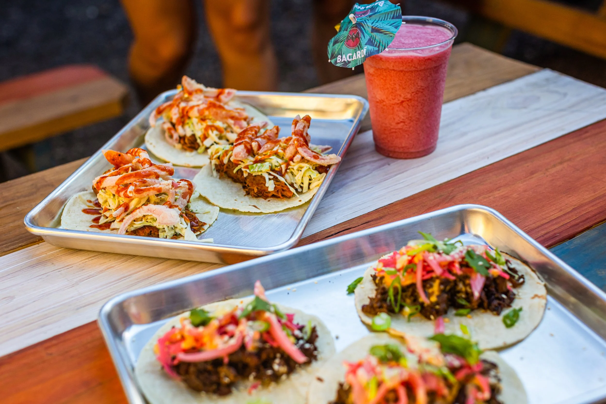 Tacos with shredded meat, vegetables, and toppings on metal trays, and a pink beverage with a decorative umbrella on a wooden table.