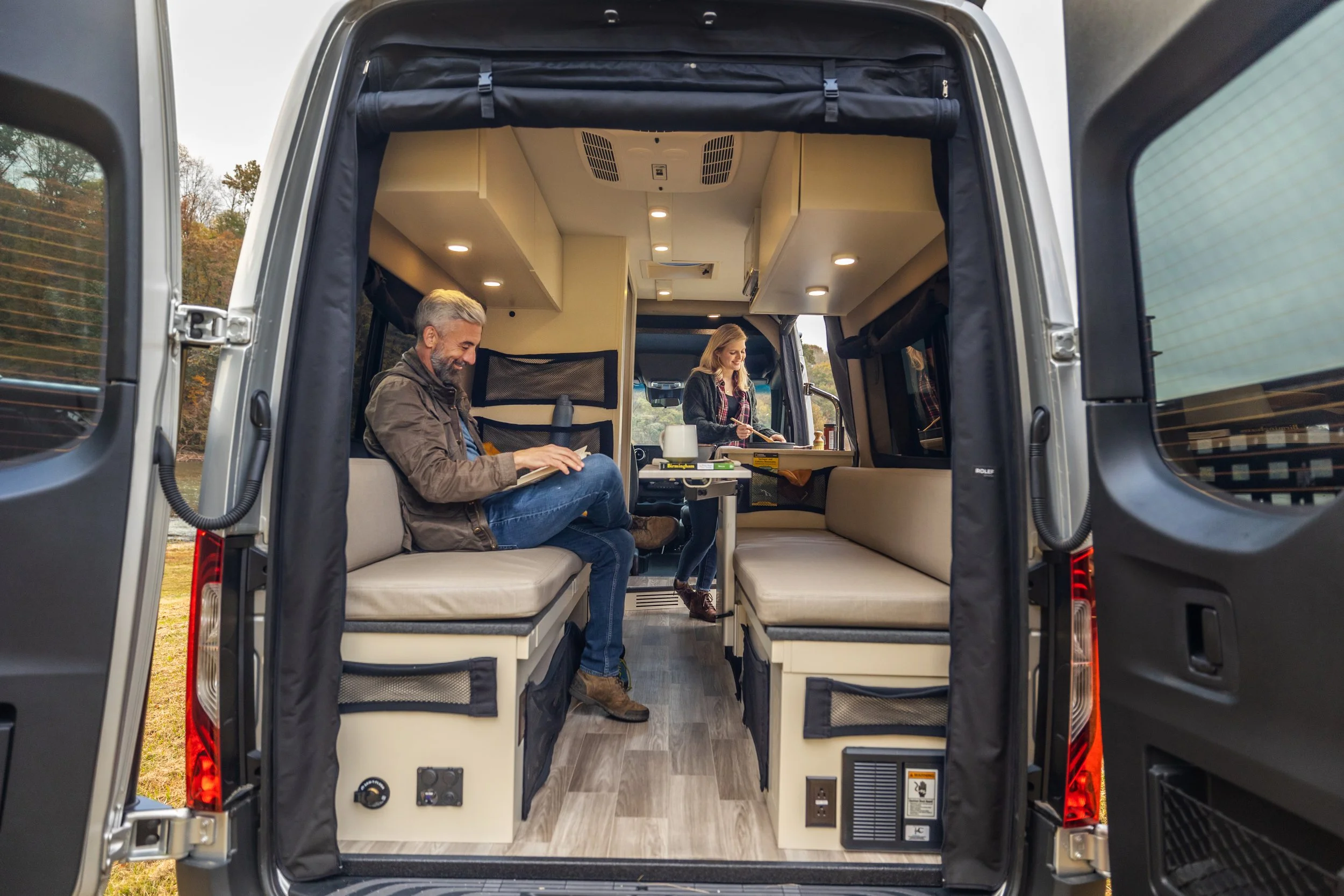Inside a compact camper van with two people relaxing; a man seated on the left, using a laptop, and a woman standing in the center preparing food at a small kitchenette.