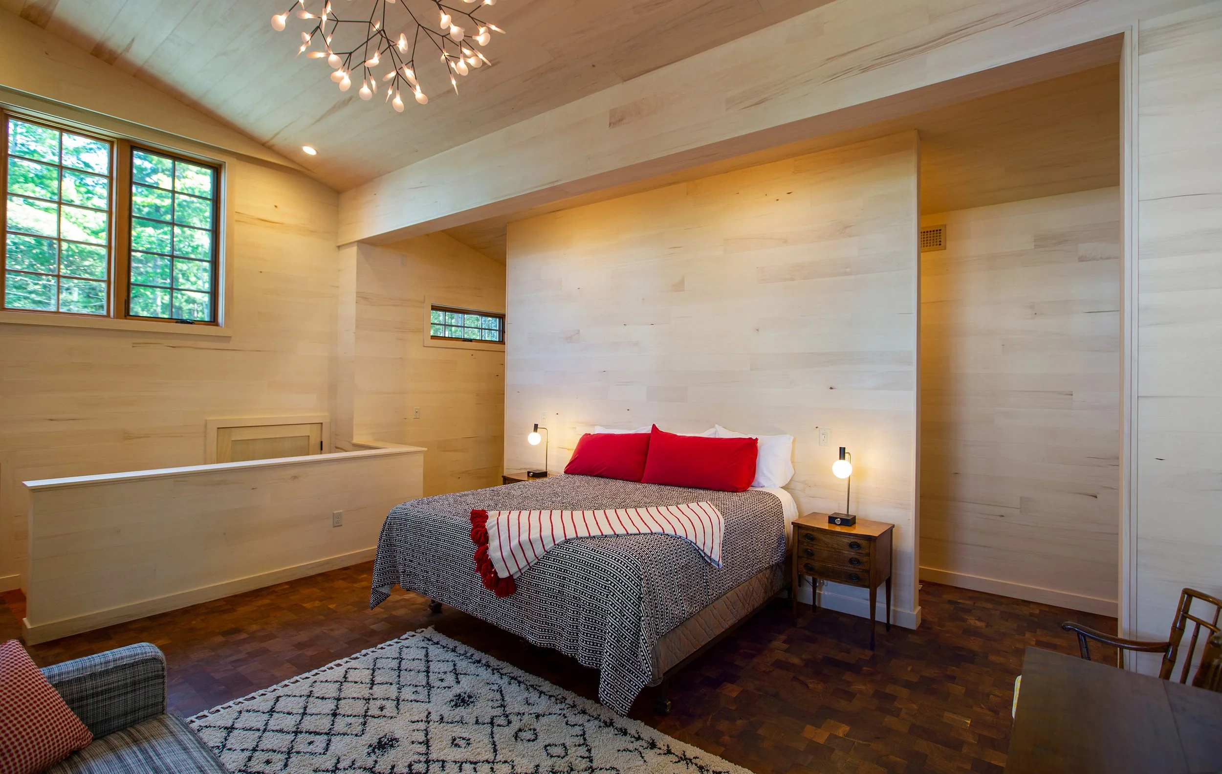 A bedroom with light wooden walls and ceiling, a large window, and a bed with red pillows and a striped blanket. There are bedside lamps, a small table, a patterned rug, and wooden furniture.