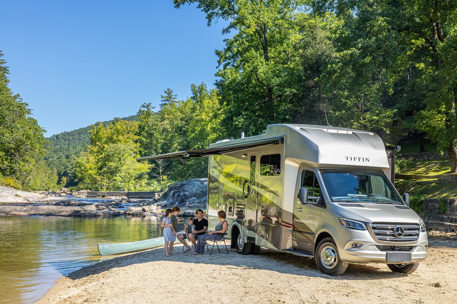 Family camping by a river with a Mercedes Tiffin RV and a canoe on the sandy shore, surrounded by green trees.