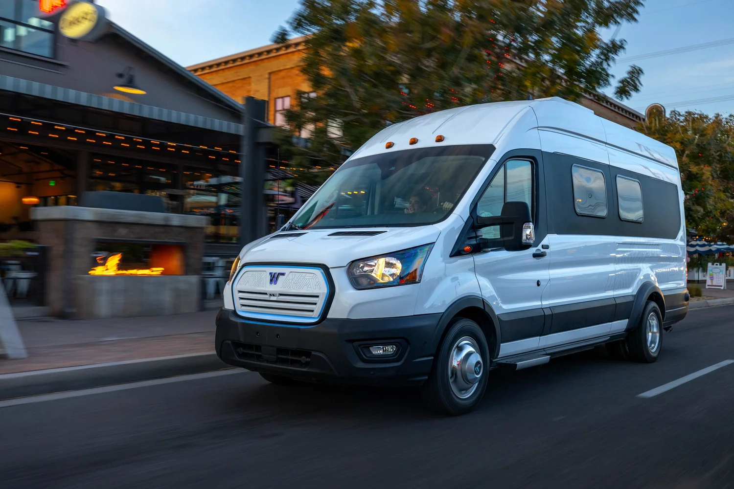 White camper van driving on a city street during early evening with blurred background of restaurants and outdoor seating