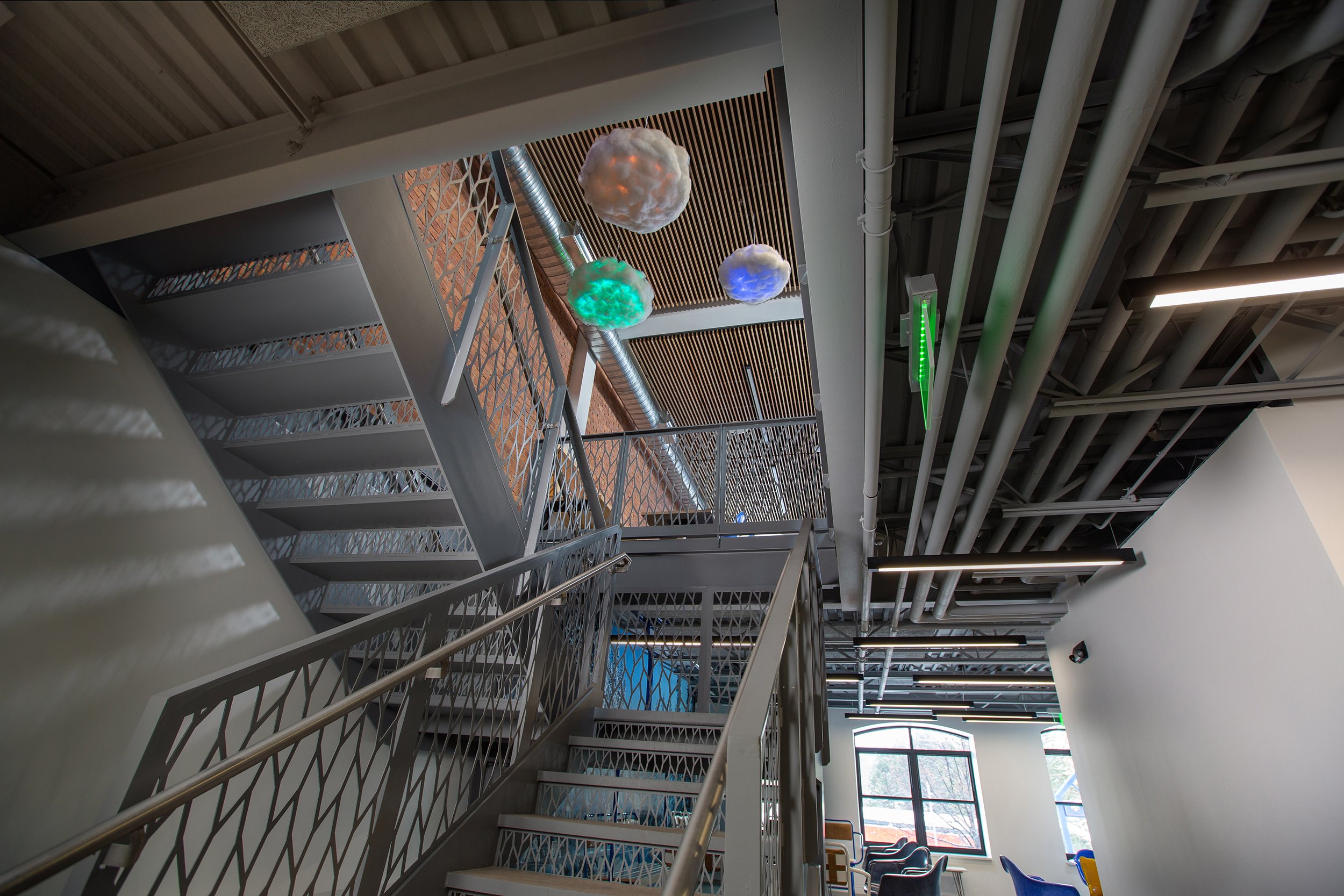 Modern indoor space with a metal staircase, colorful cloud-shaped light fixtures hanging from the ceiling, and large windows letting in natural light.