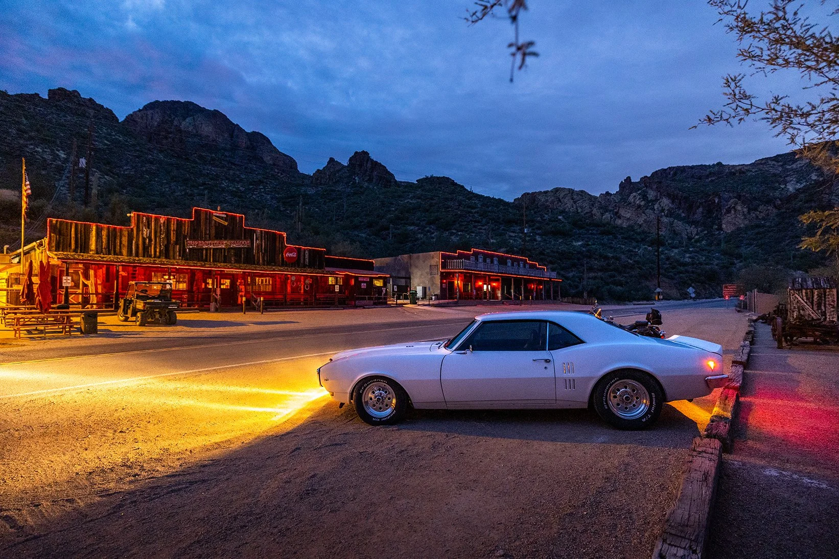 A classic silver car parked on the side of a deserted street at dusk, with a mountain range and old western-style buildings illuminated by red neon lights in the background.