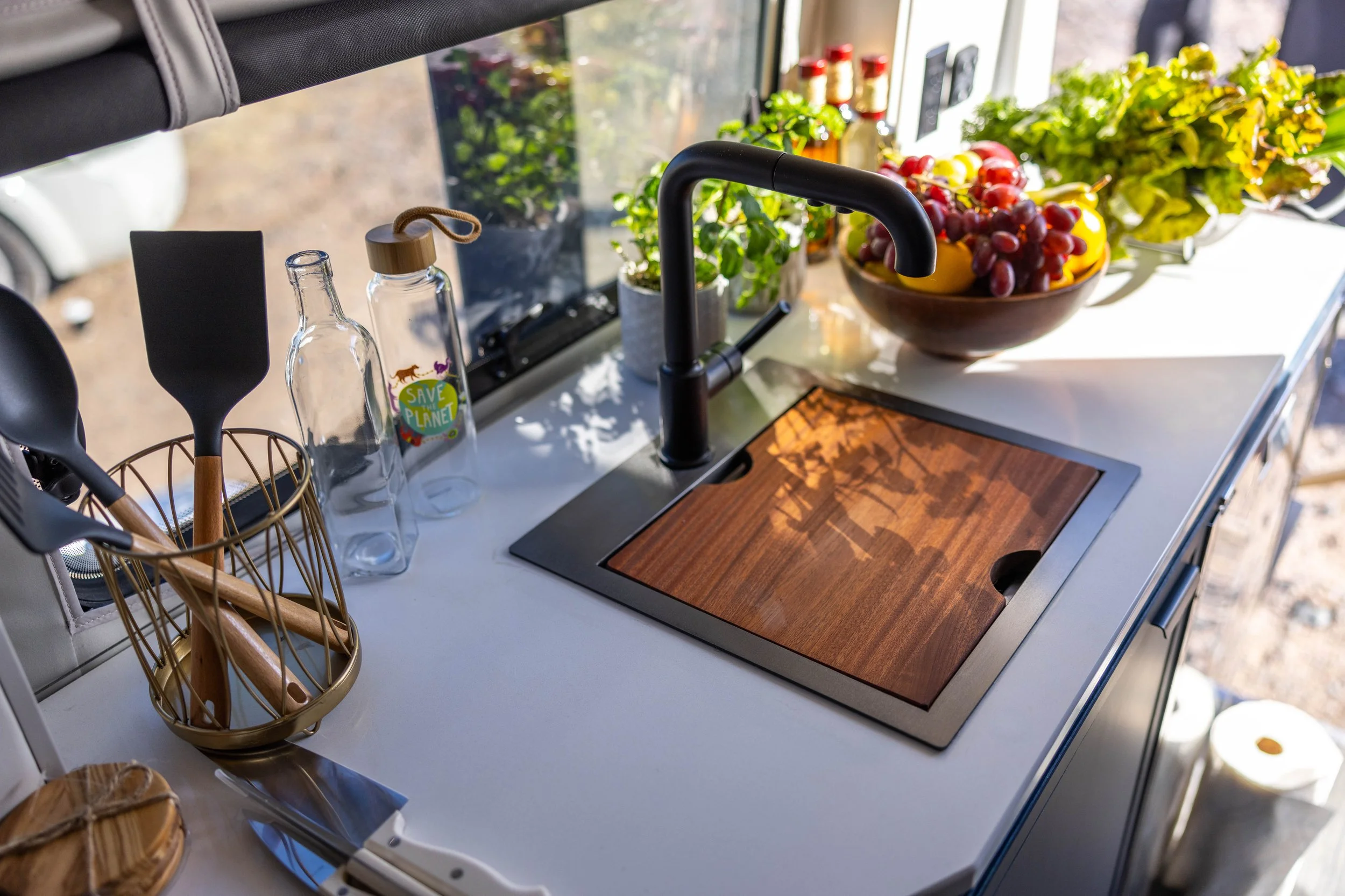 Kitchen countertop with a black sink, a wooden cutting board, a bowl of fresh grapes and yellow peppers, bottles, and utensils.