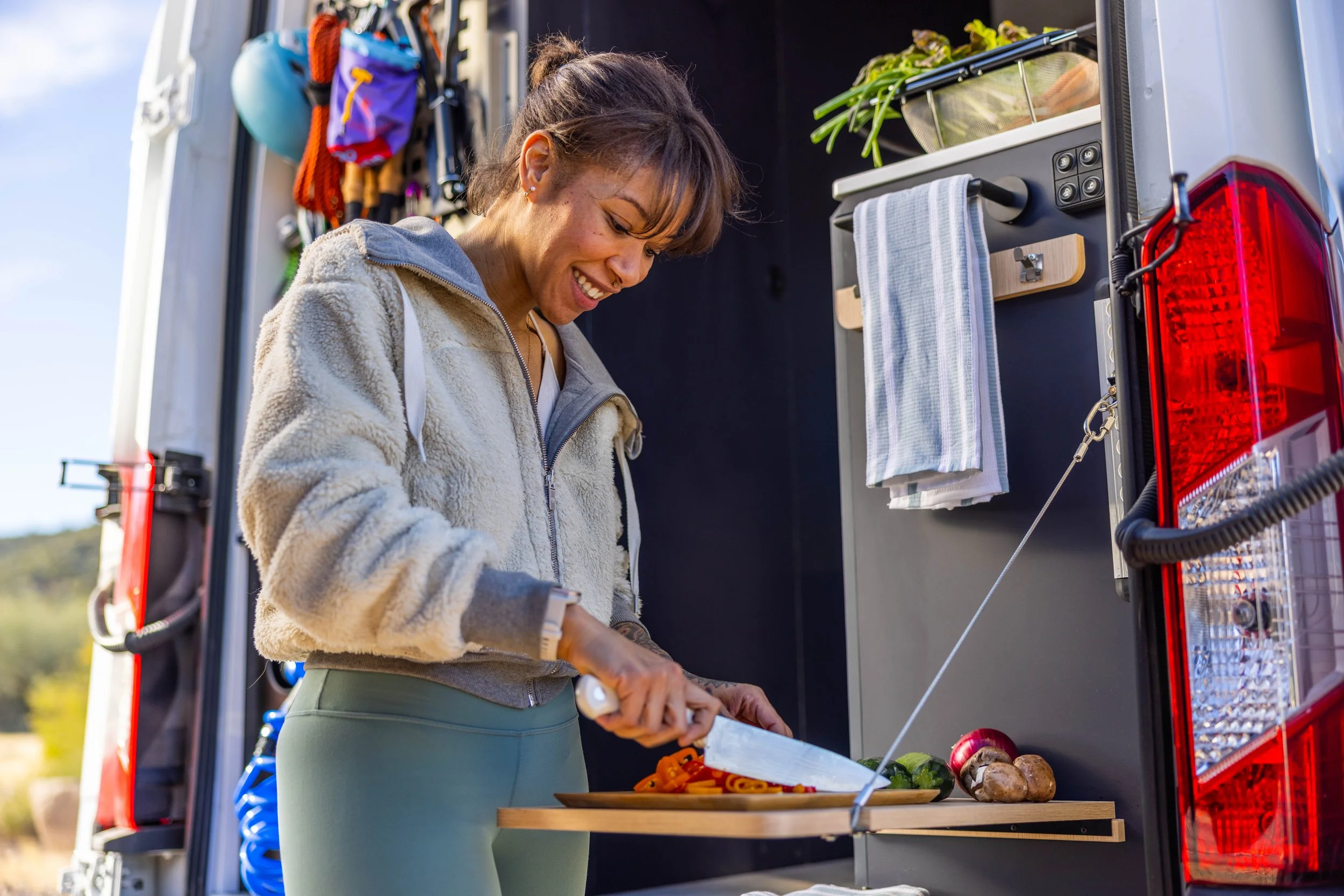 A woman is smiling and cooking food with vegetables and pasta in a mobile kitchen or food truck, outdoors on a sunny day.