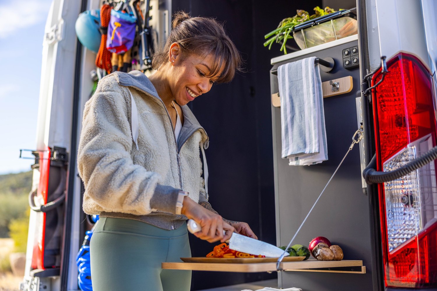 A woman is smiling and cooking food with vegetables and pasta in a mobile kitchen or food truck, outdoors on a sunny day.