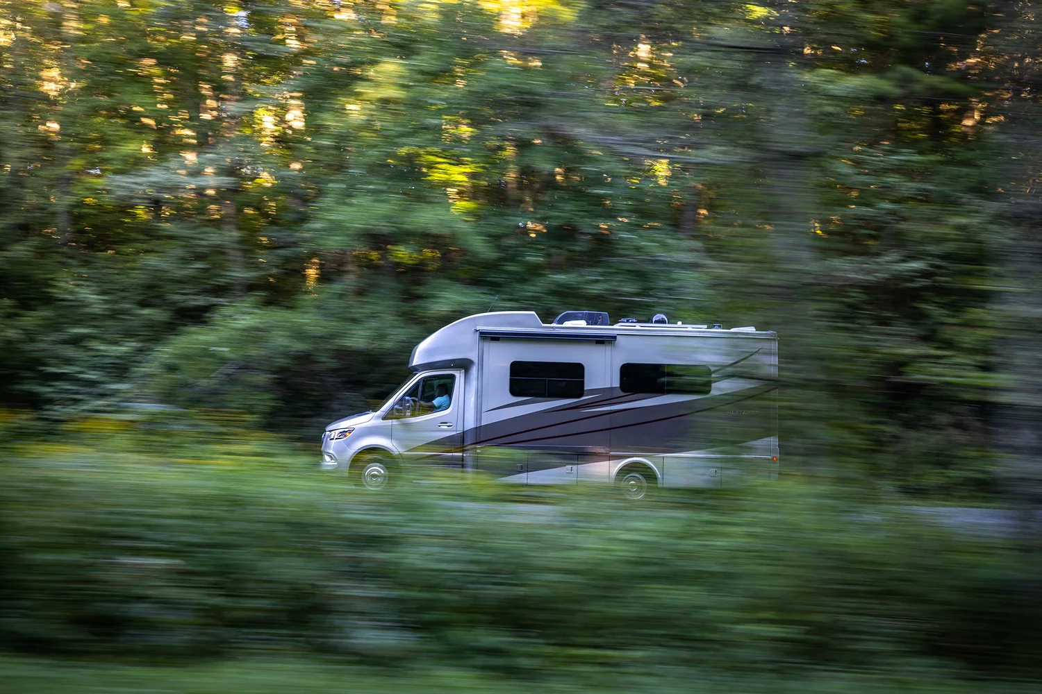 A silver-colored recreational vehicle (RV) driving through a lush green forest with motion blur, indicating movement.