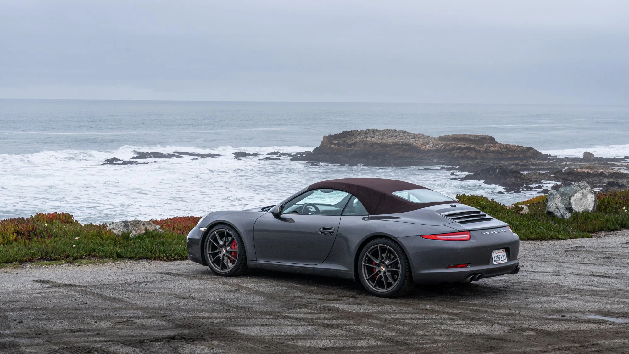 Gray Porsche convertible parked on a coastal road with waves and rocks in the background
