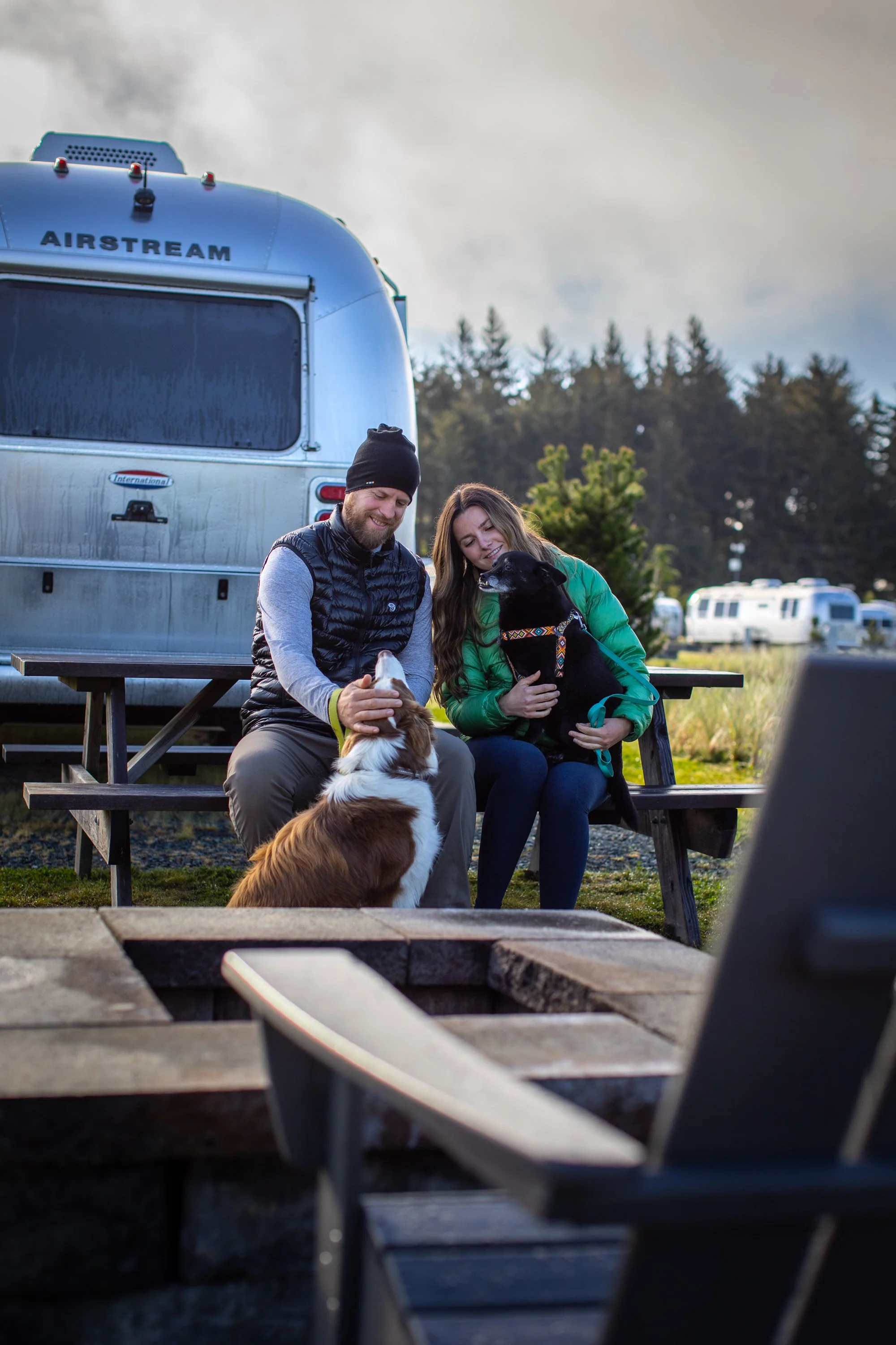 Two people sitting on a picnic bench outdoors, each with a dog, in front of an Airstream trailer, with trees and other trailers in the background, during daytime.