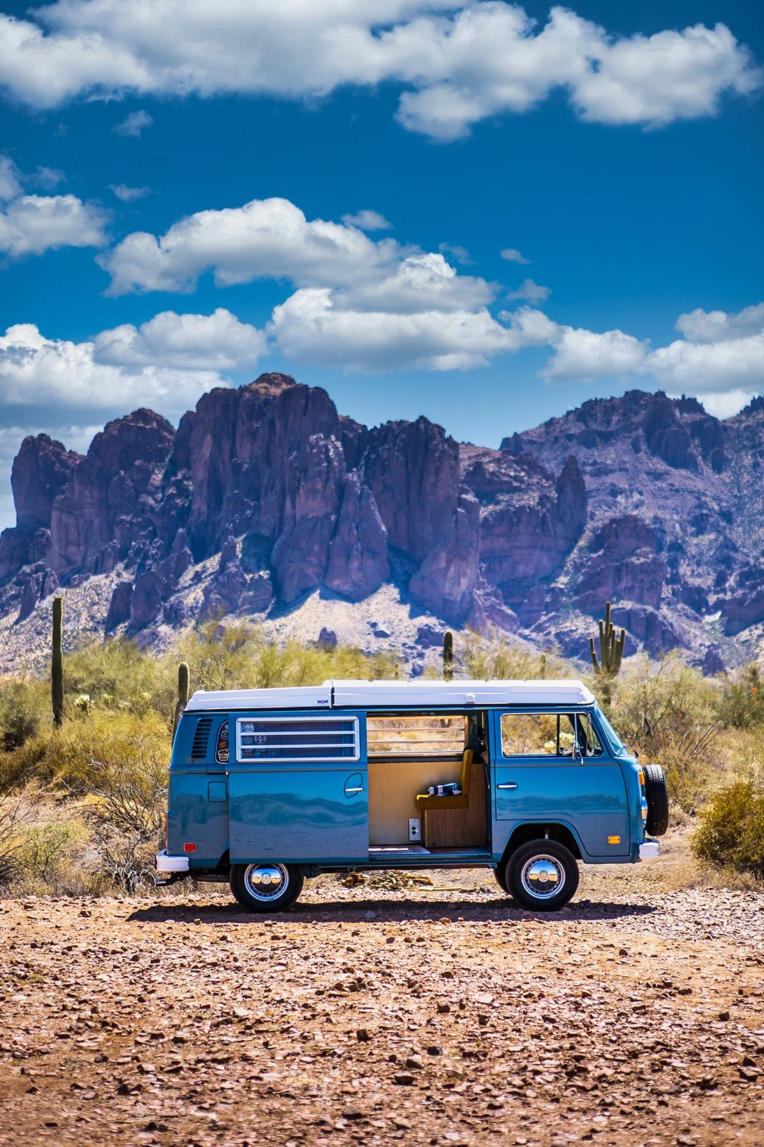 A vintage blue camper van parked on a dirt desert road with cacti and shrubs. Behind are rugged mountains under a partly cloudy sky.