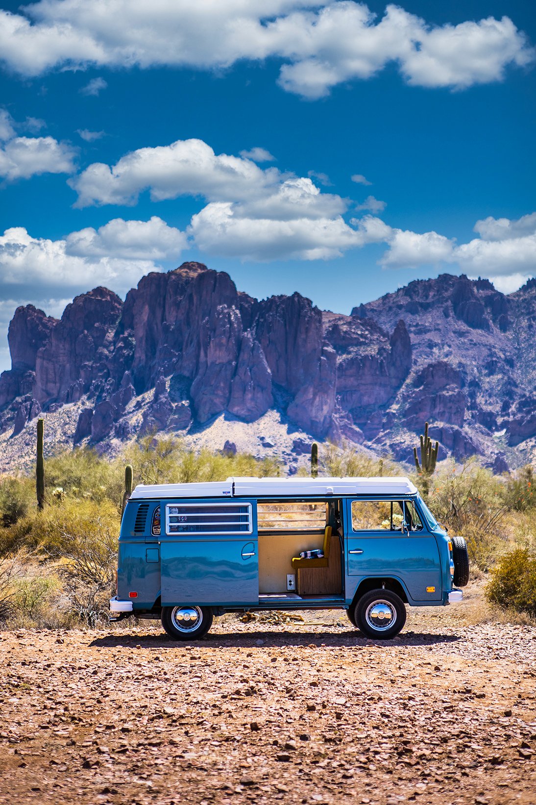 A vintage blue camper van parked on a dirt desert road with cacti and shrubs. Behind are rugged mountains under a partly cloudy sky.