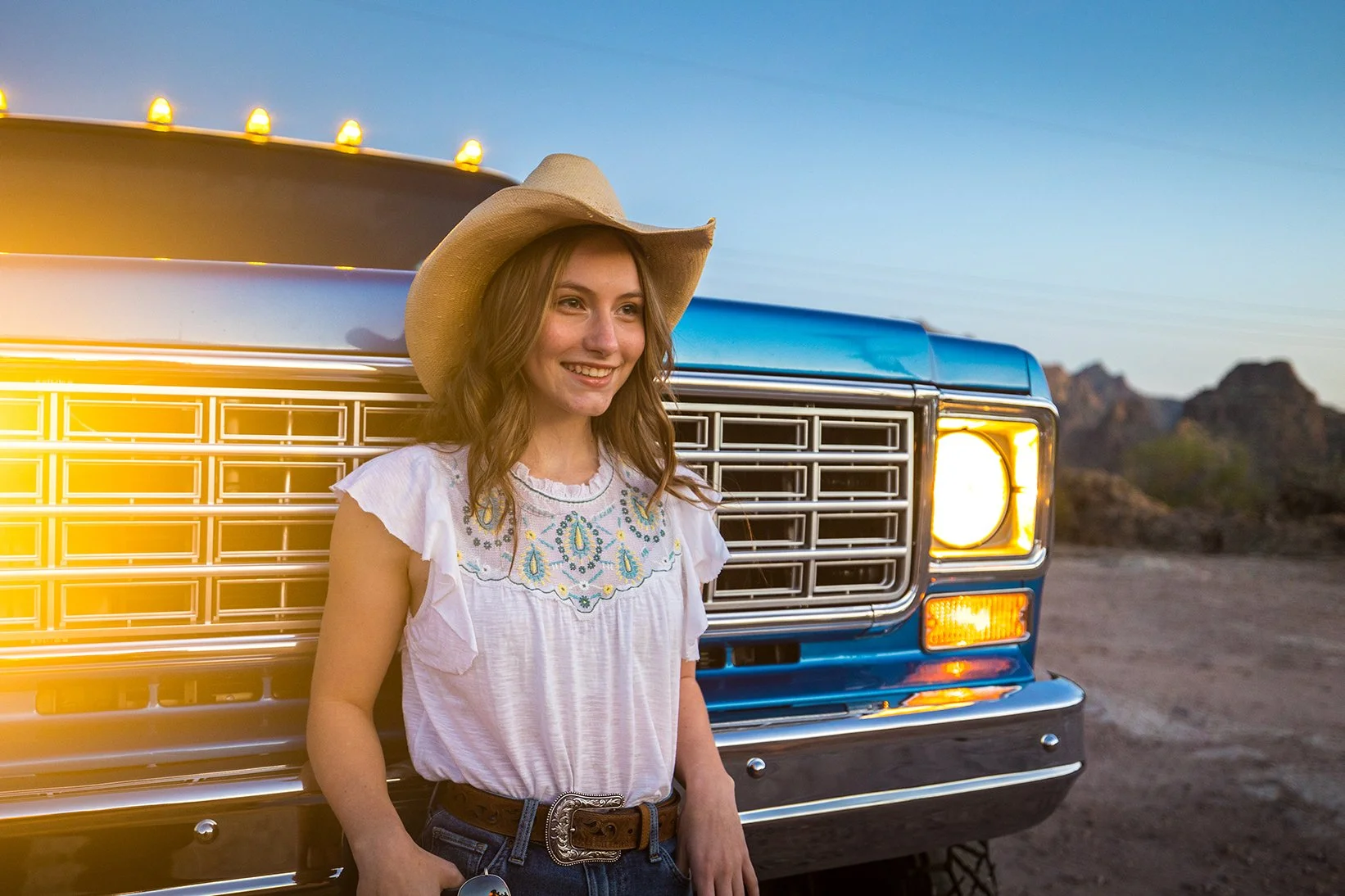 A young woman with wavy red hair wearing a cowboy hat, white embroidered blouse, and jeans, smiling and leaning against a vintage blue truck in a desert landscape during sunset.