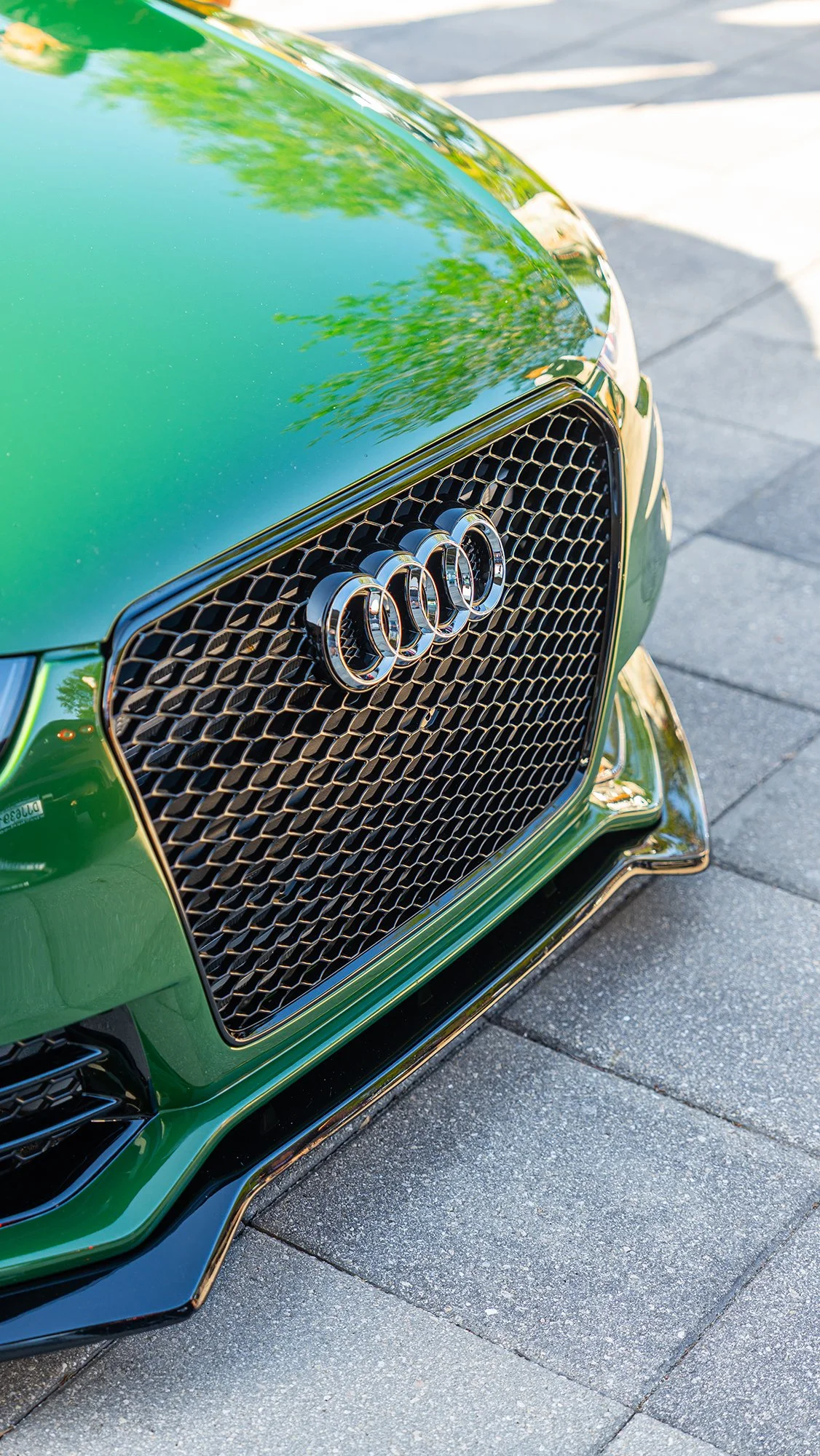 Close-up of a green Audi car front grille with the Audi logo, parked on a paved street.