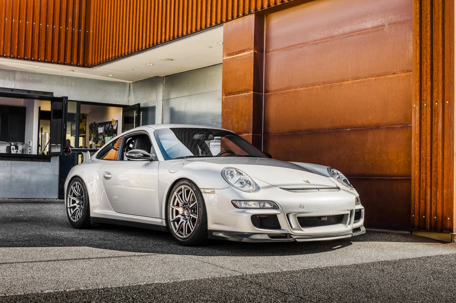 Silver Porsche sports car parked outside a modern building with wooden and concrete exterior.
