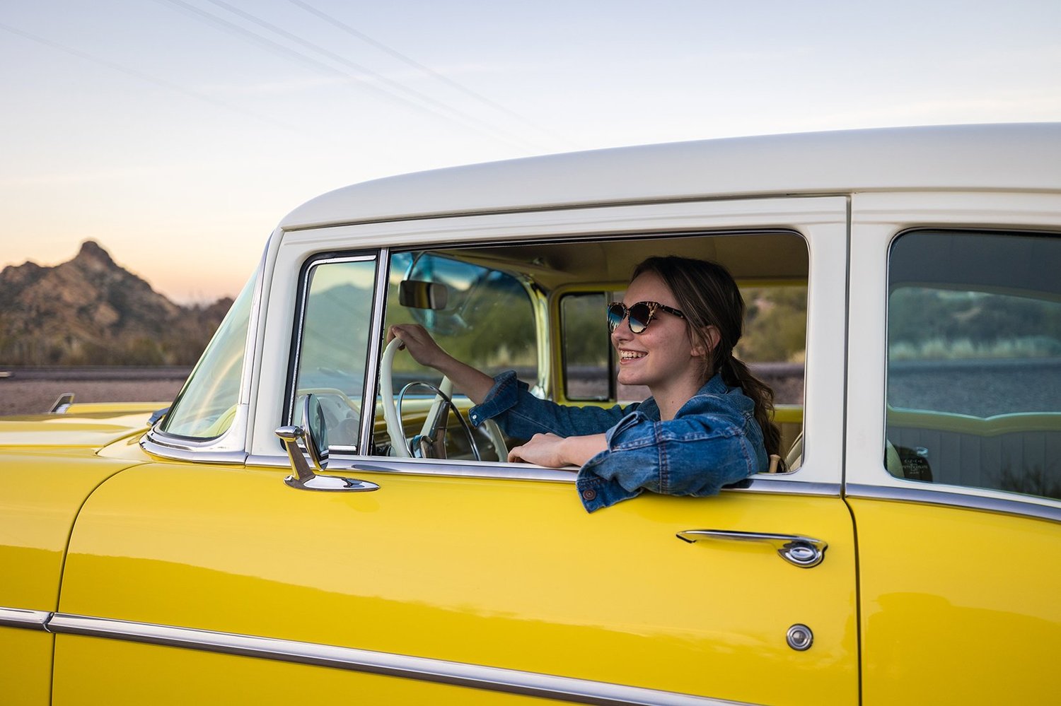 Smiling woman in sunglasses and denim jacket sitting in the driver's seat of a yellow vintage car with a desert landscape and mountains in the background.