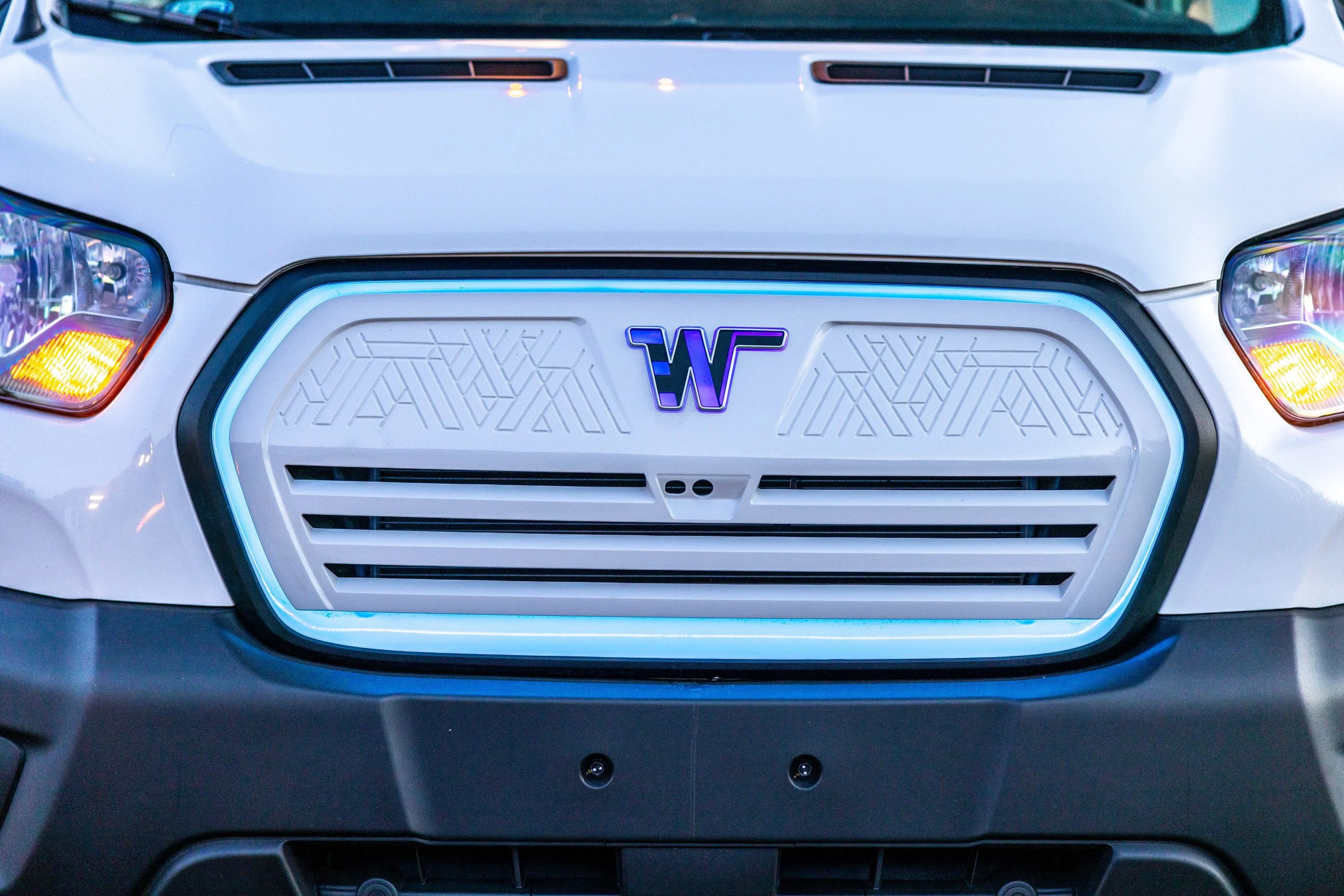 Front view of a white vehicle with a futuristic grille and illuminated blue light accent, featuring a purple and black 'W' logo in the center.